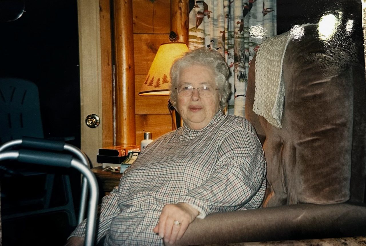 An elderly woman sits on a brown recliner in a cozy living room beside a lit lamp.