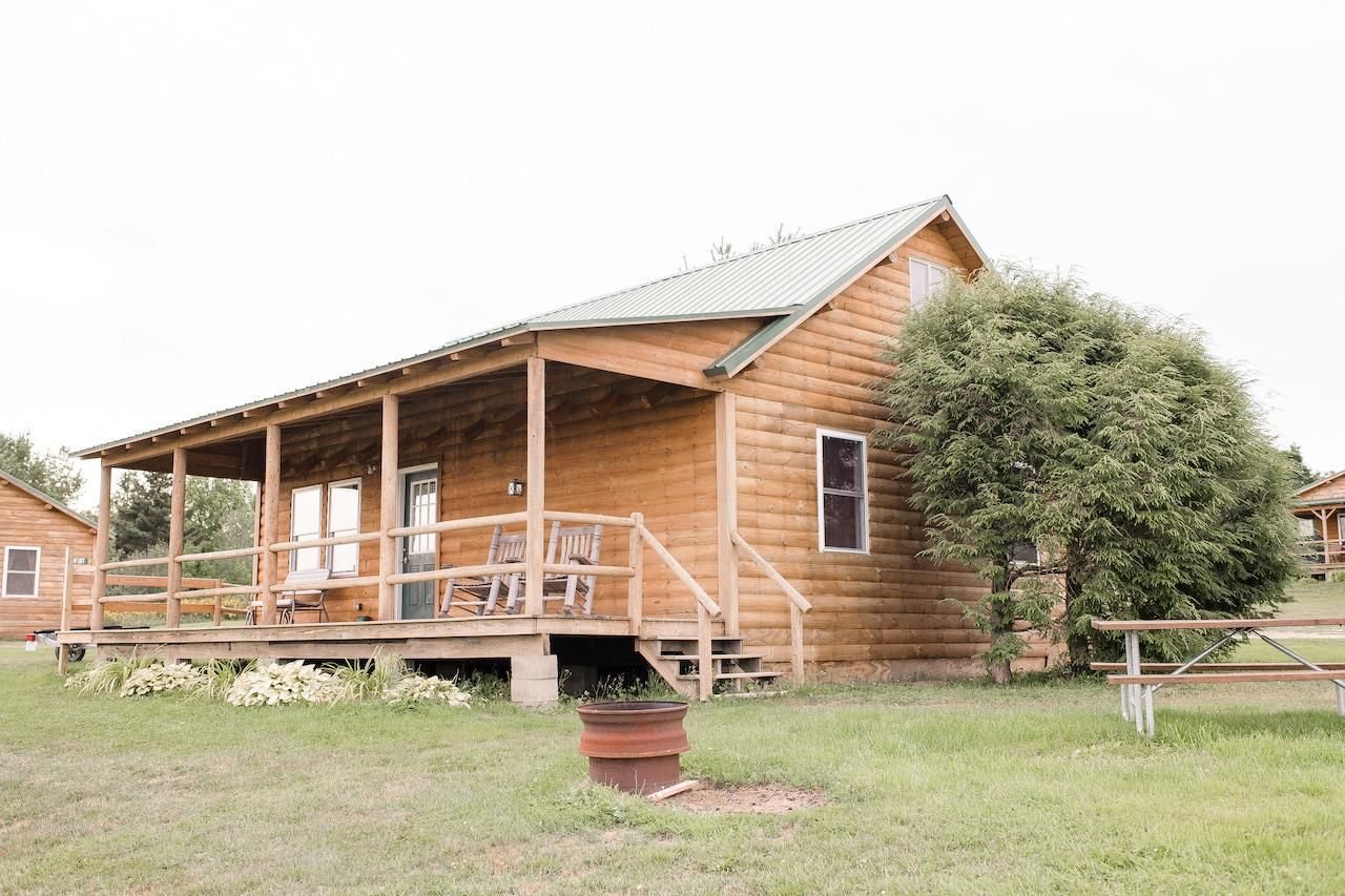Rustic wooden cabin with a porch, in a grassy yard beside a tree and picnic table.