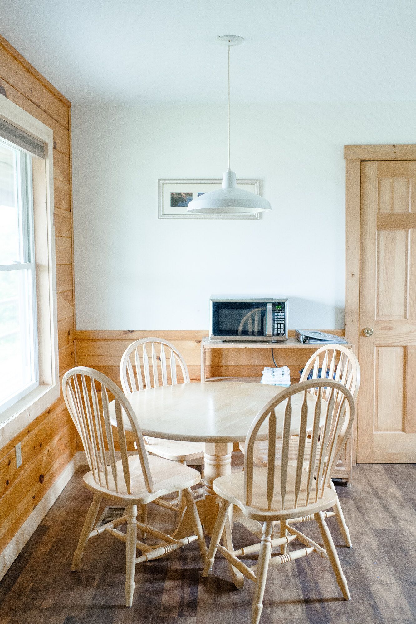 Bright dining room with a round wooden table, four chairs, a microwave, and wood paneling.
