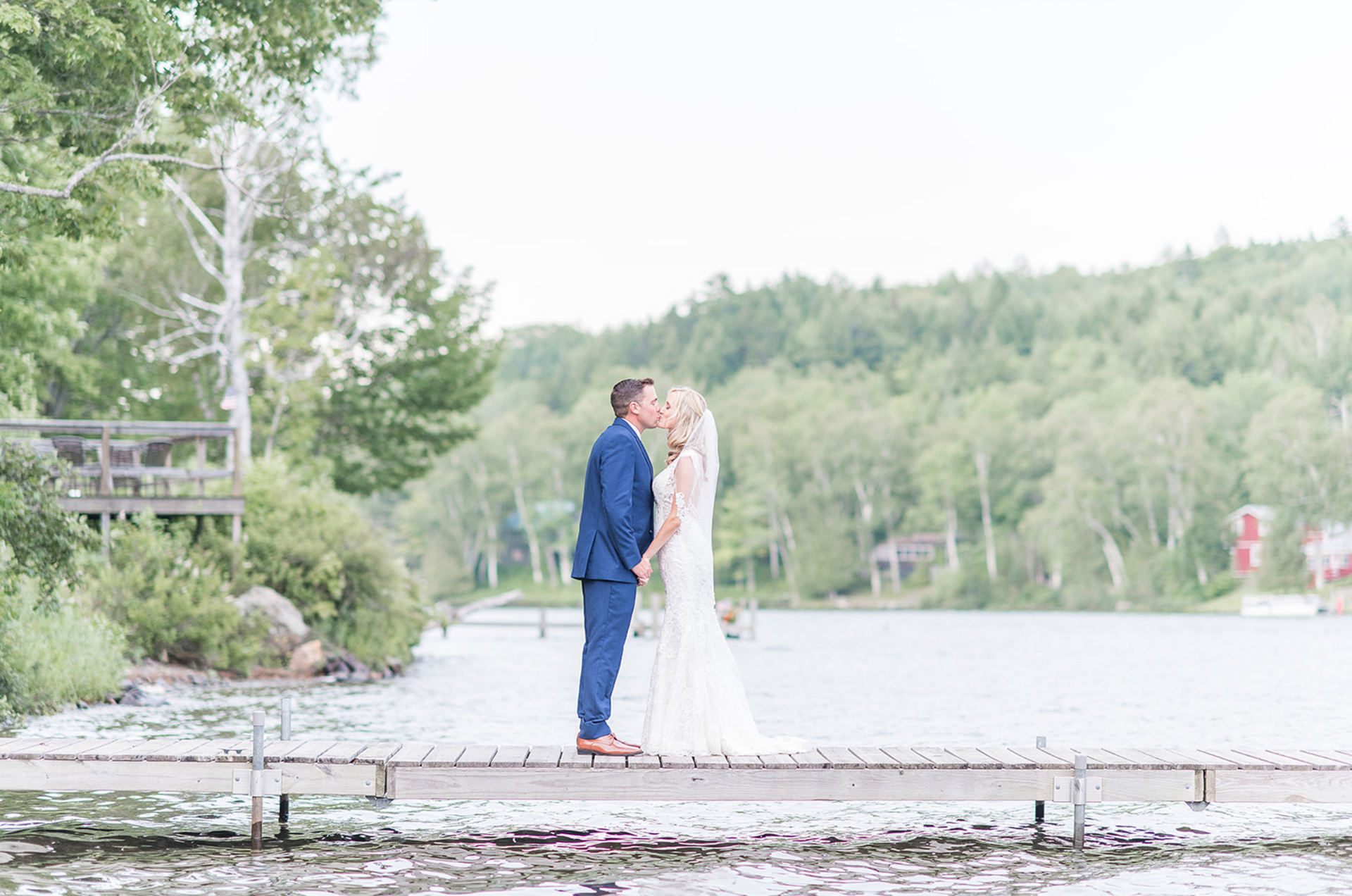 Couple in blue suit and white dress kissing on a lakeside dock with forested hills in the background