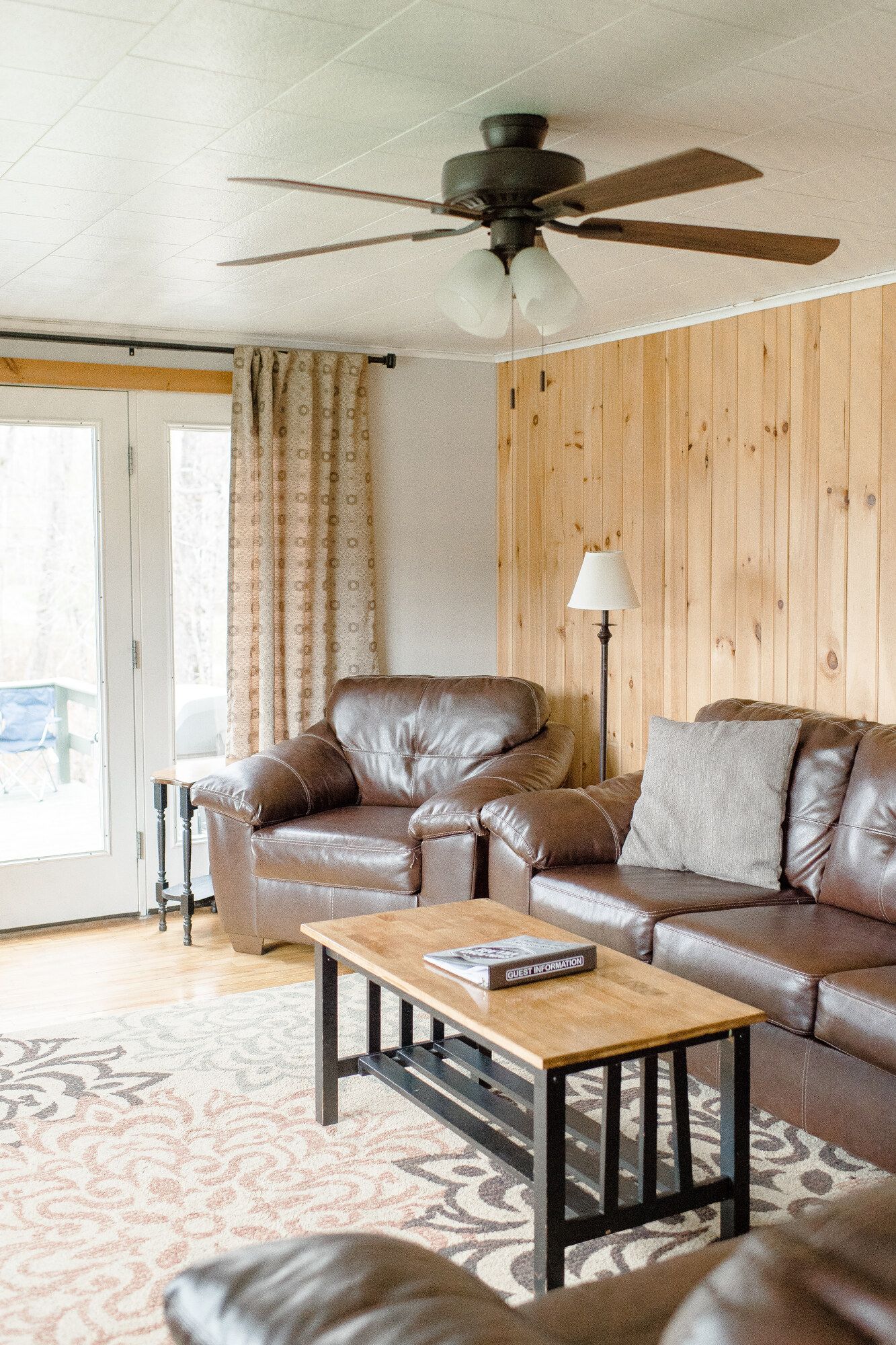 Living room with brown leather sofa, coffee table, floor lamp, wood-paneled wall, and ceiling fan