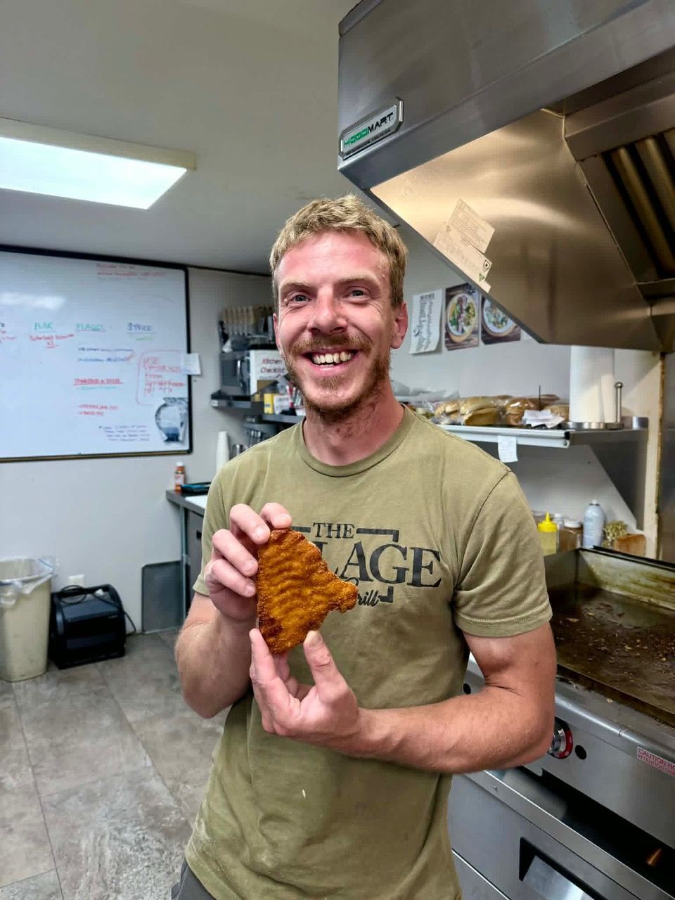 Smiling man in a kitchen holding a crispy fried item near a stainless steel stovetop.