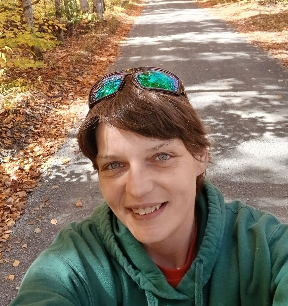 Selfie on a tree-lined road in autumn, with a smiling person wearing sunglasses on their head and a green top