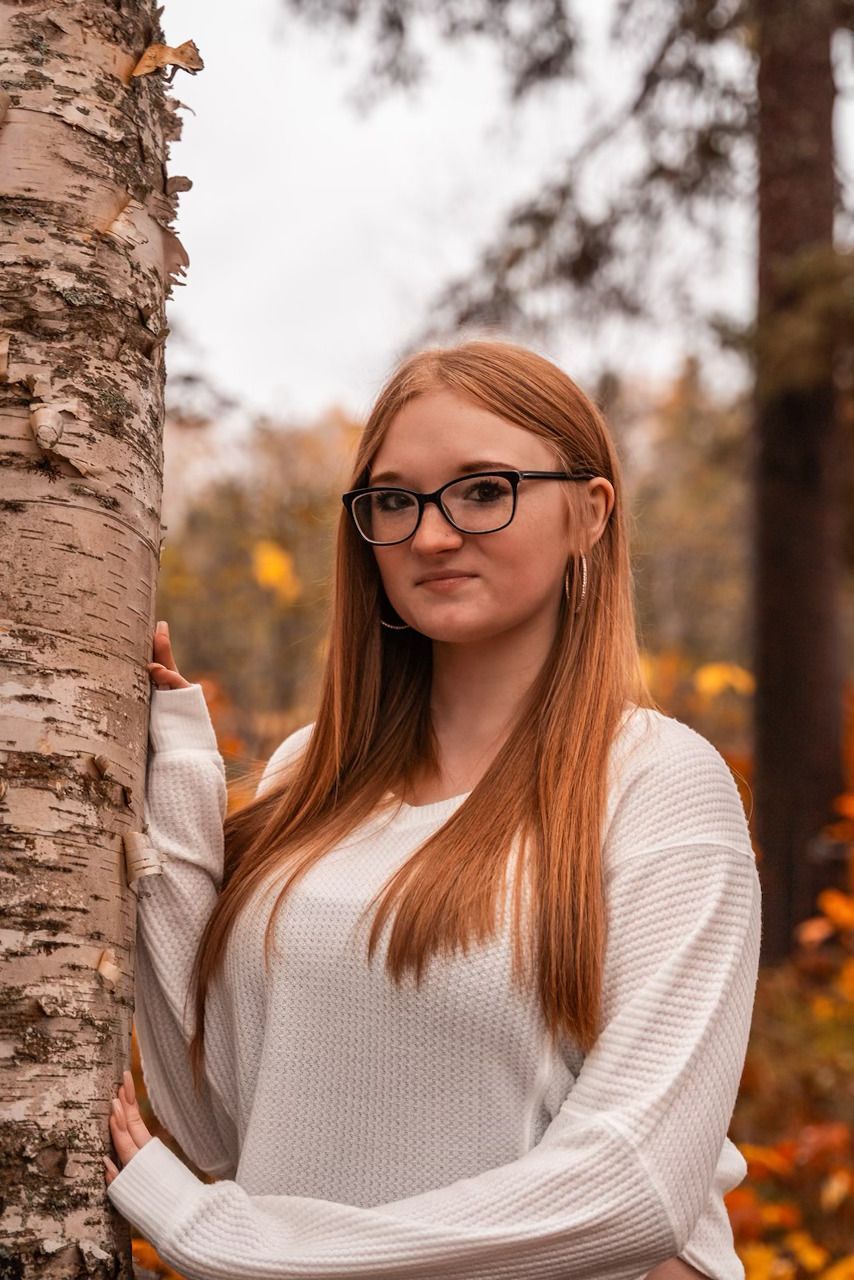 Woman with long red hair and glasses standing by a birch tree in an autumn forest