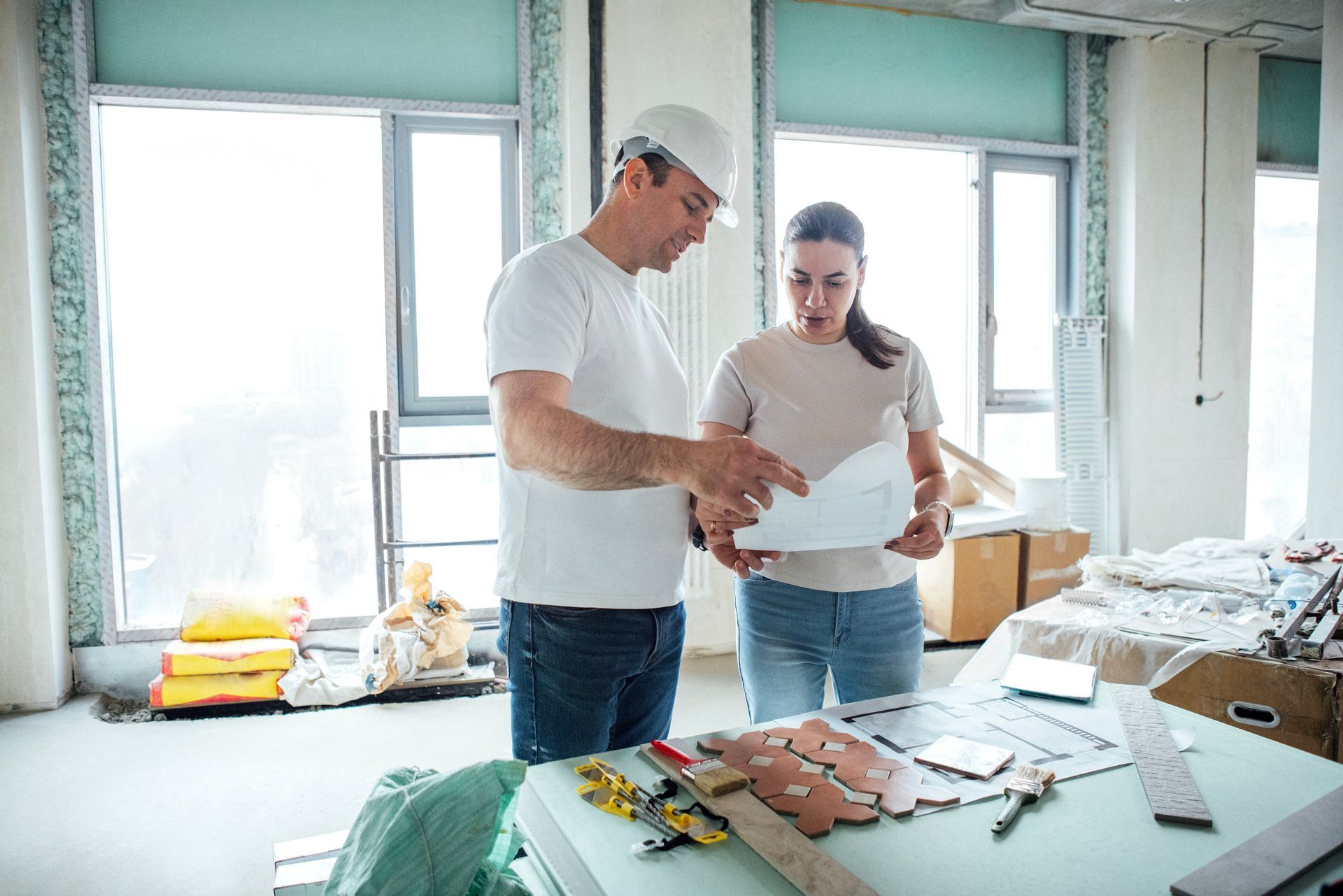 Man in hard hat and woman reviewing blueprints in a construction site with tools on a table.