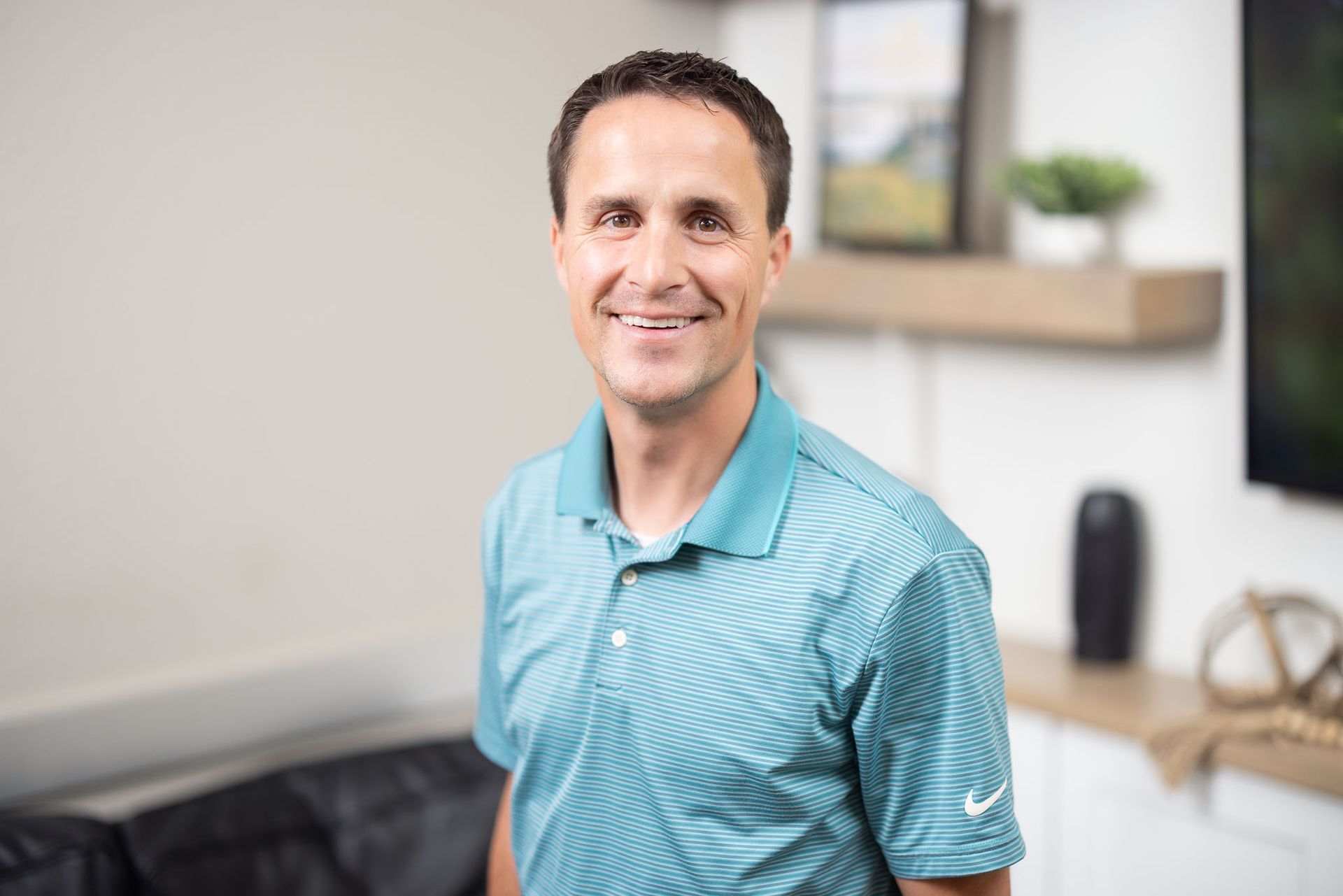 Taylor Wade smiling in a teal polo shirt in a bright office, standing beside an exam table