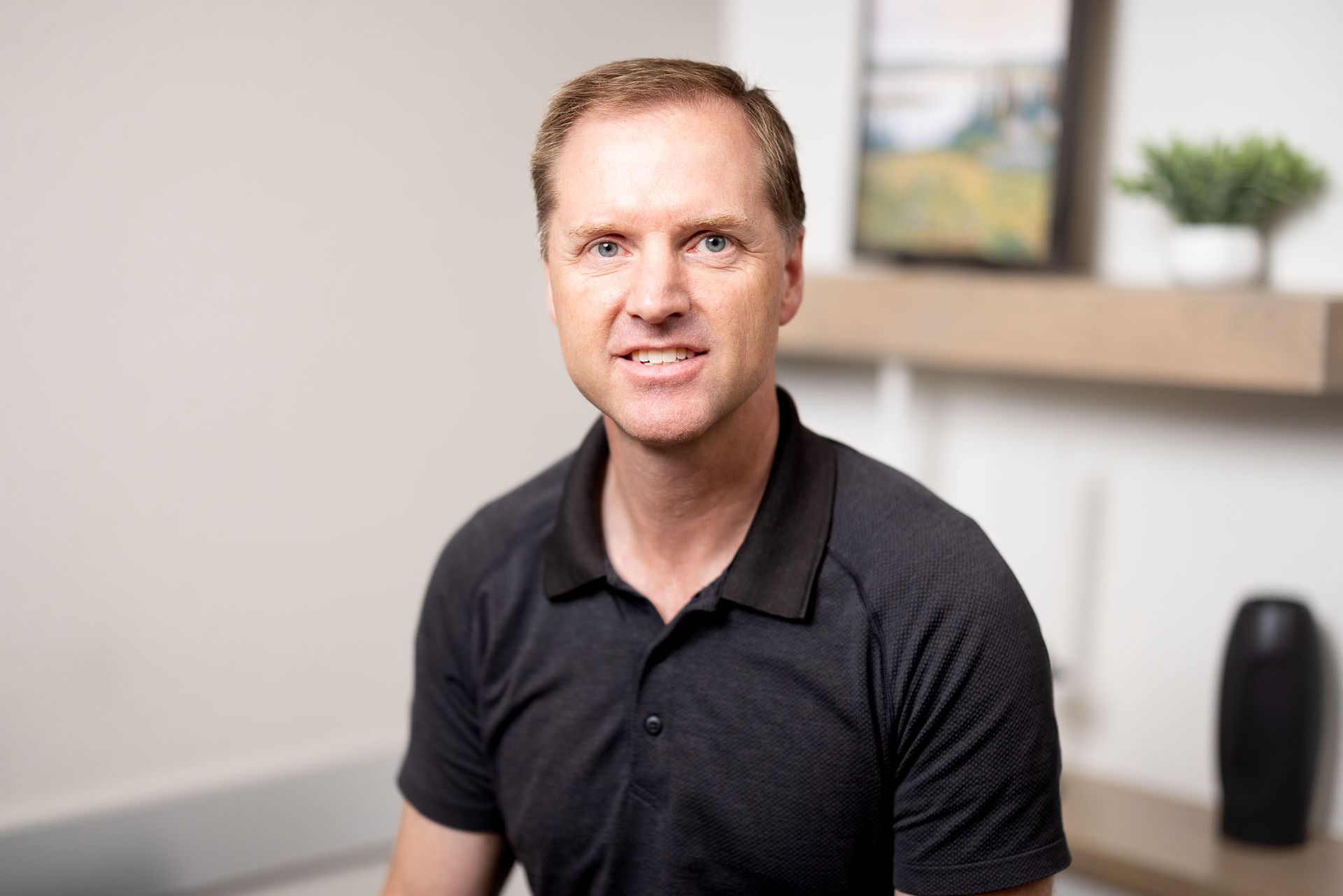 Lance Harmon, MD in a black polo shirt seated indoors, with a shelf and plant in the background.