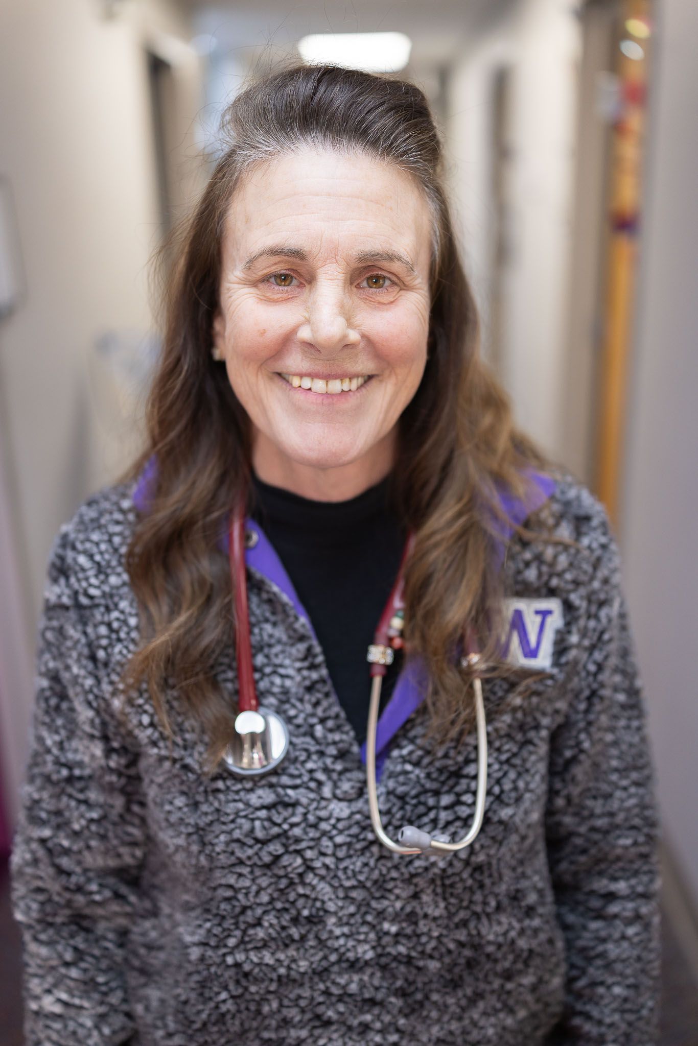 Smiling healthcare worker in gray sweater with stethoscope, standing in a hospital hallway.