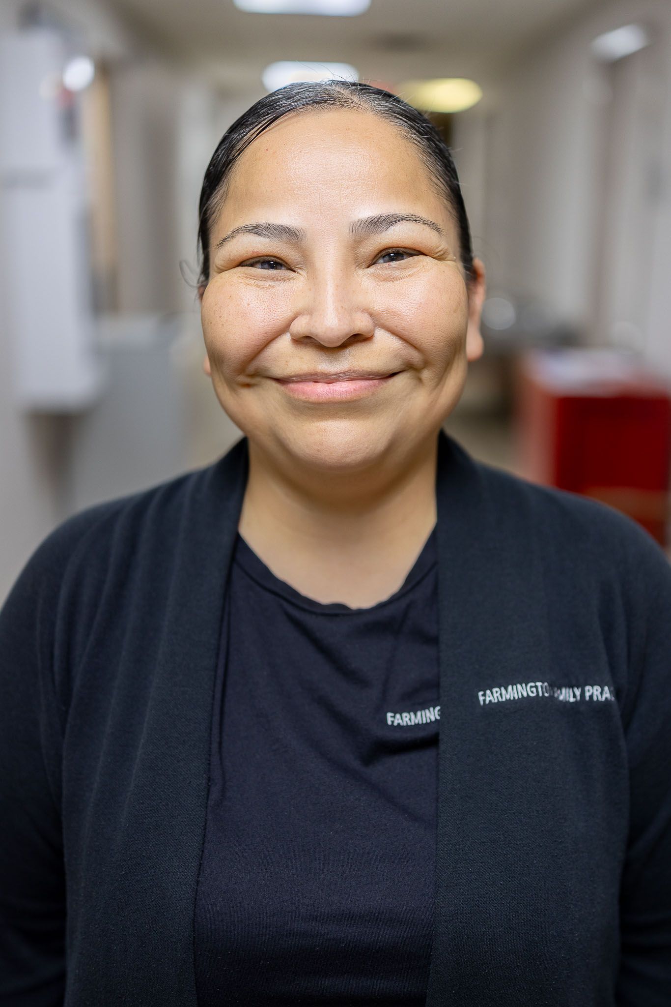 Smiling Farmington Family Practice employee in a black top standing in a softly blurred hallway
