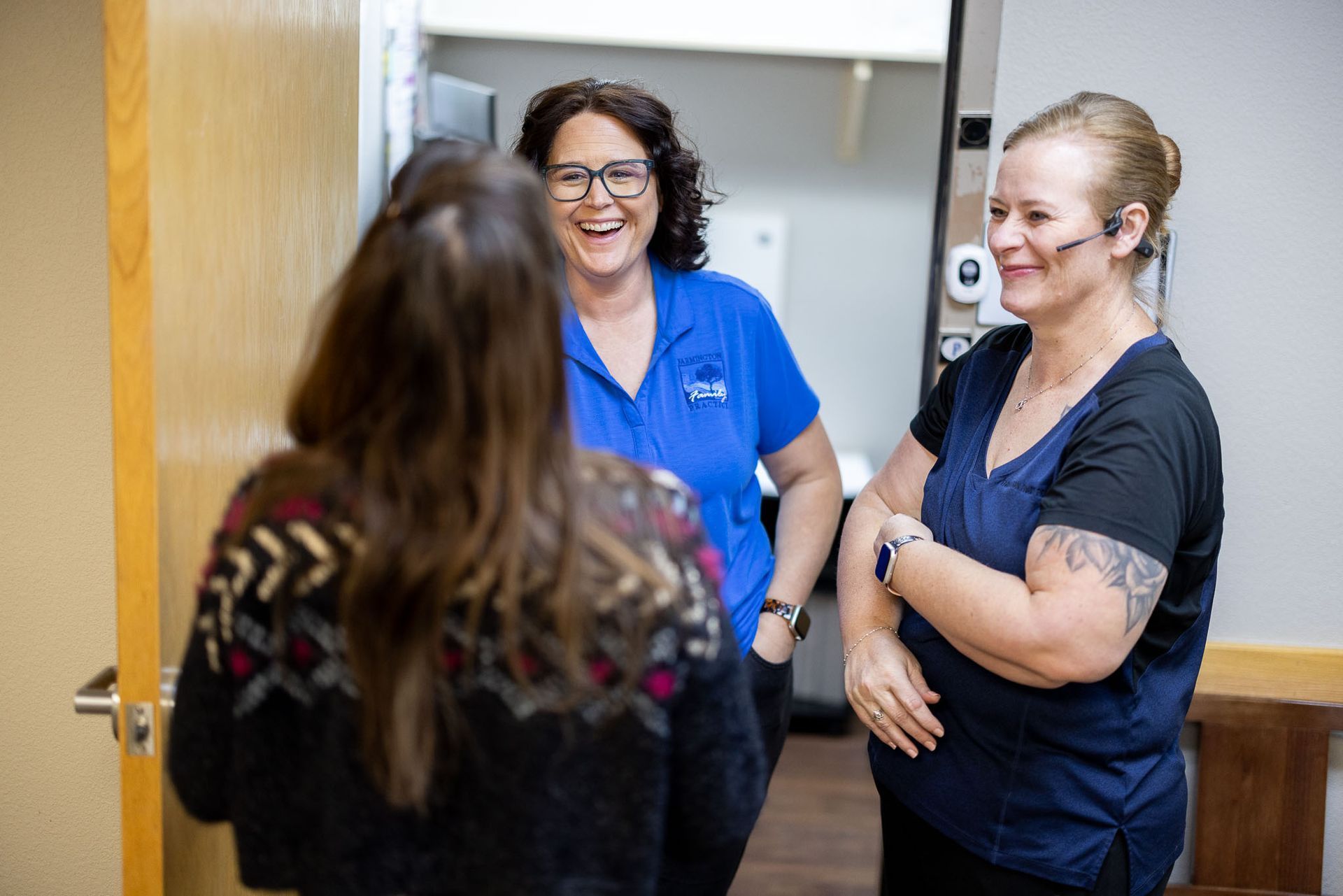 Three people talking in a hallway, two smiling nurses in blue scrubs greeting a visitor near an open door.