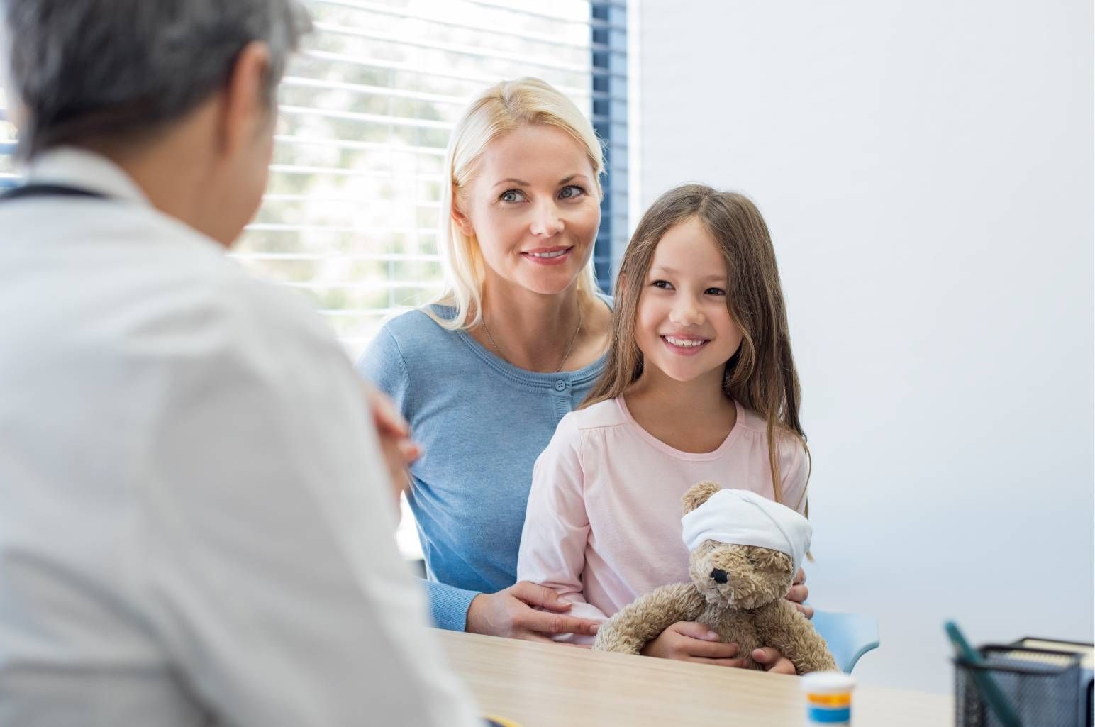 Woman and girl smiling at a doctor in a bright clinic, holding a small stuffed toy on the exam table