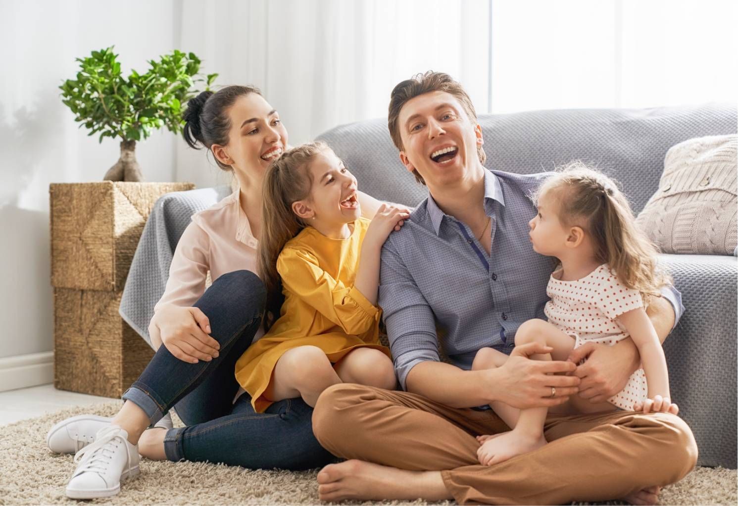 Family laughing together on a living room floor with a couch and potted plant behind them