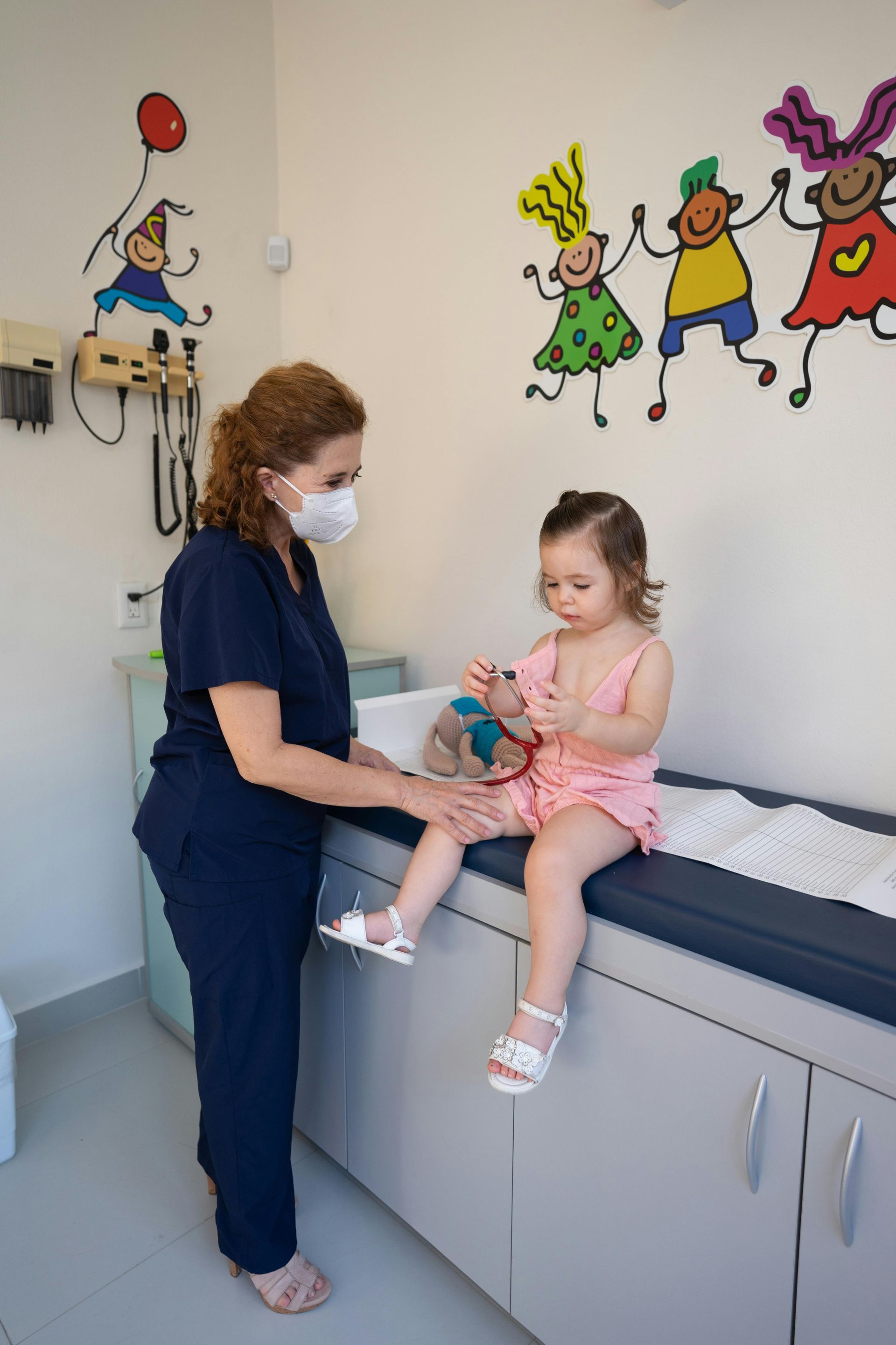Child sitting on an exam table while a masked nurse stands beside her in a pediatric clinic room