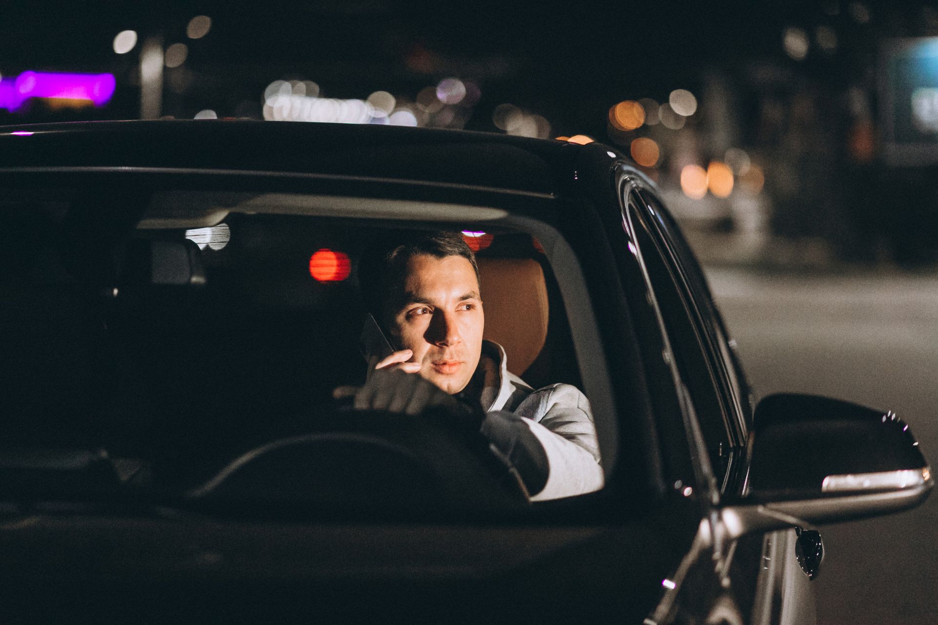 Man in car at night, smoking, looking thoughtful. Dark setting with city lights.