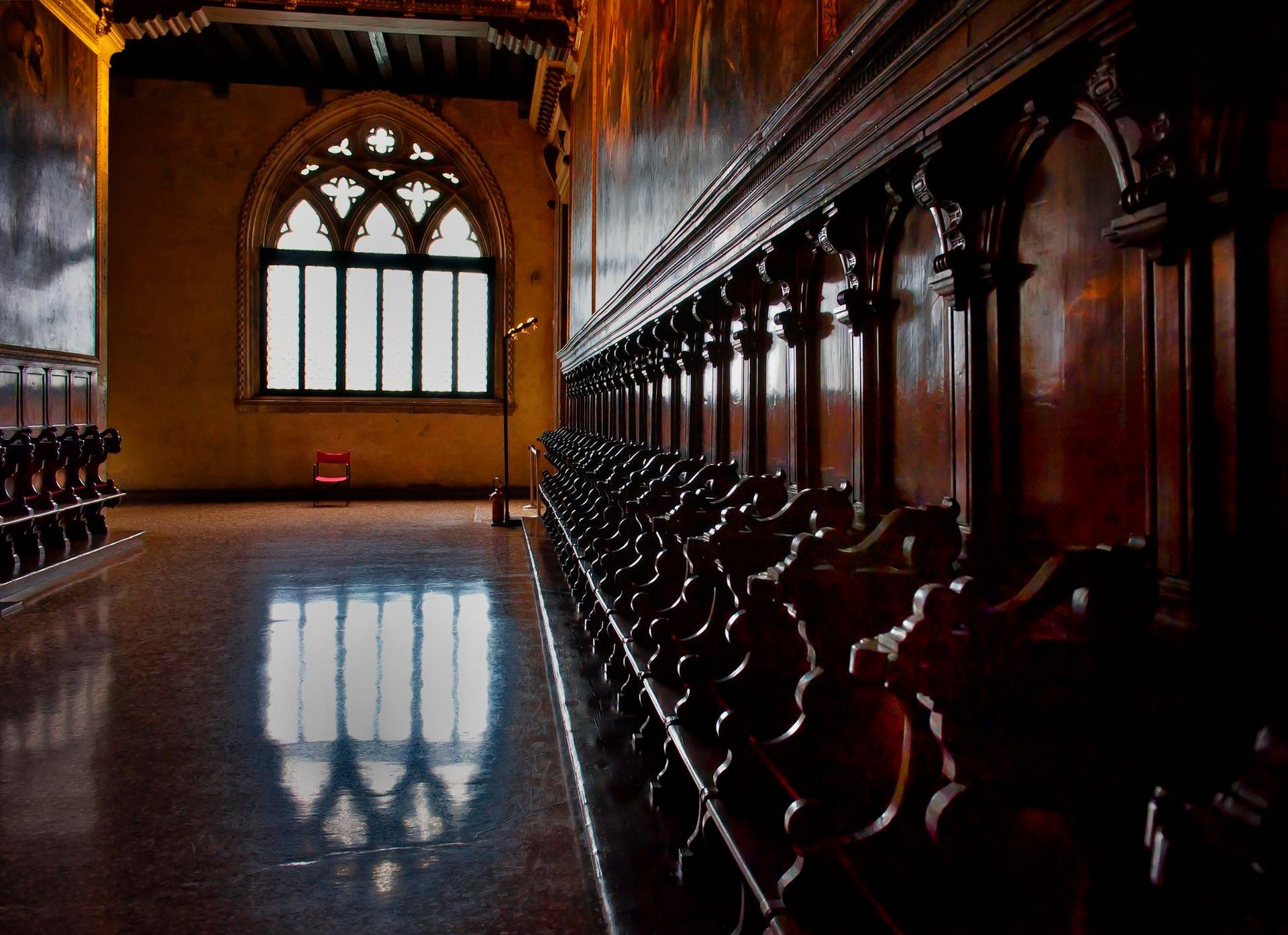 Dark, ornate wooden choir stalls in a church. Gothic window reflects light on polished floor.