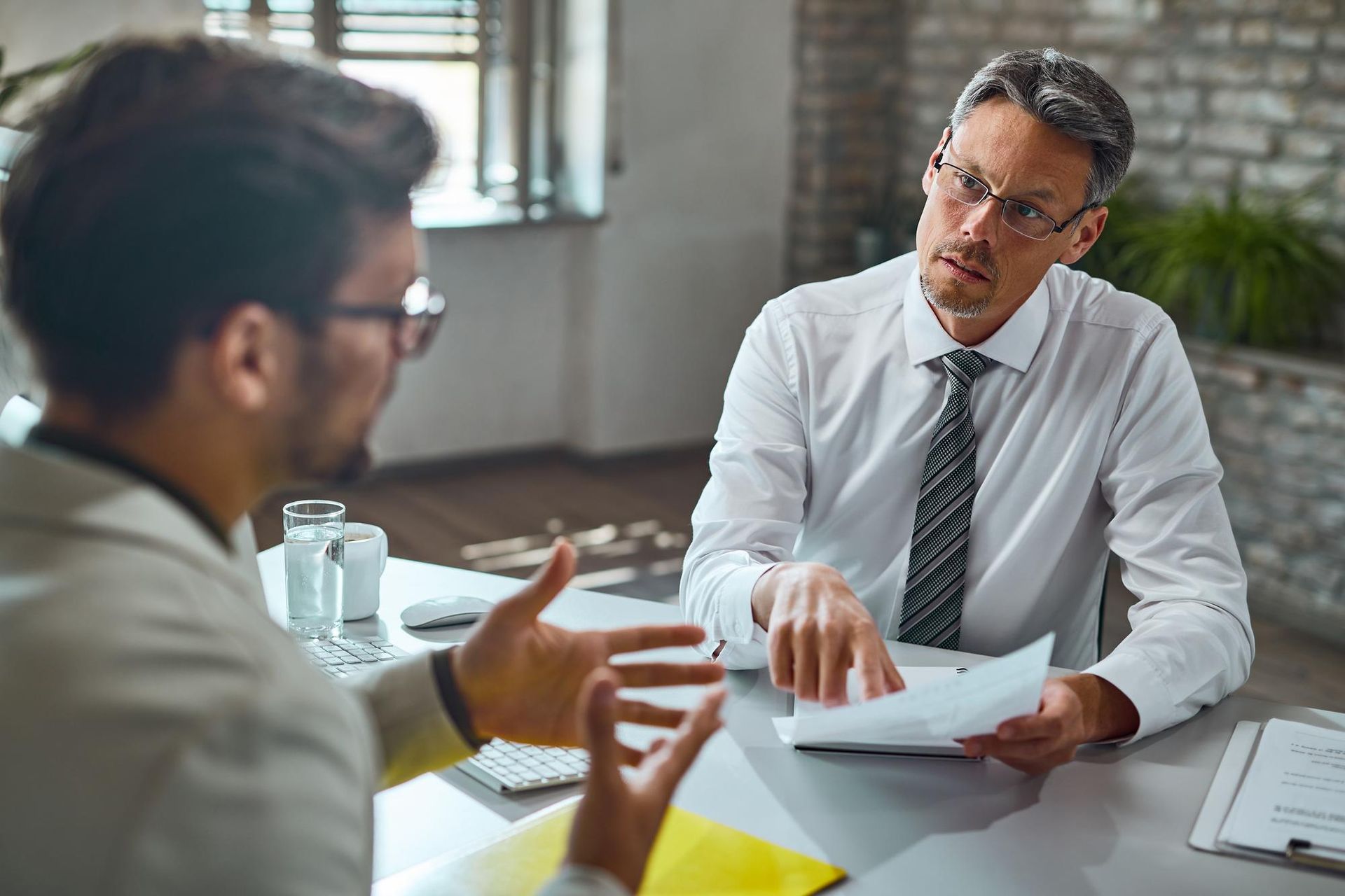 Two men in an office setting; one gestures while talking, the other looks at papers.
