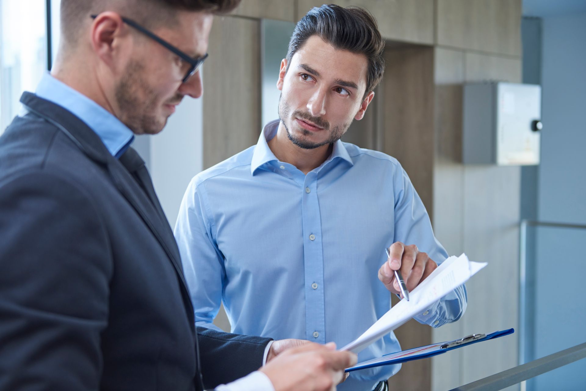 Two men in office setting reviewing paperwork; one pointing, the other looking on.