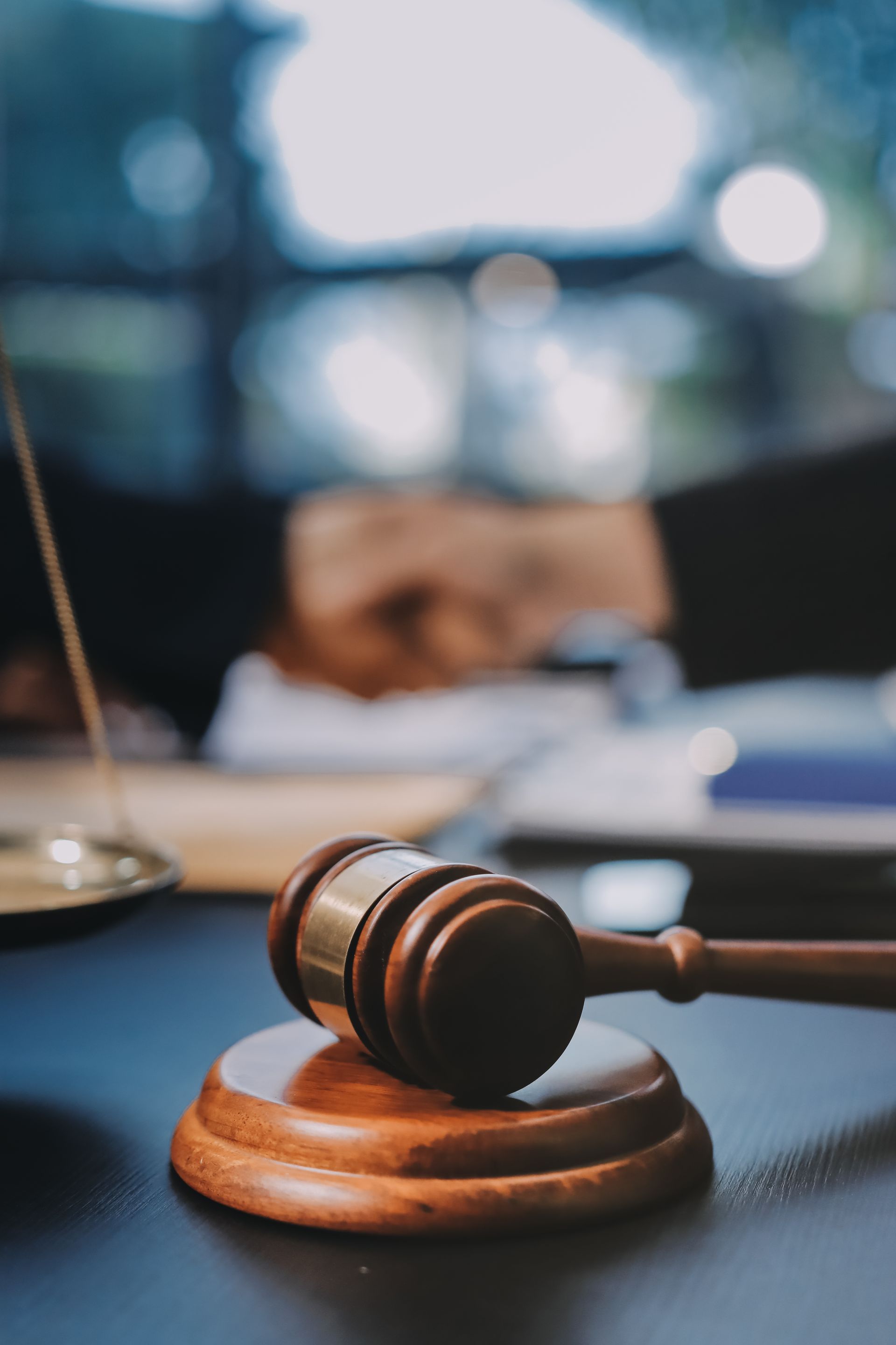 Gavel on wooden block on a dark surface, scales of justice in the background, blurred office setting. 