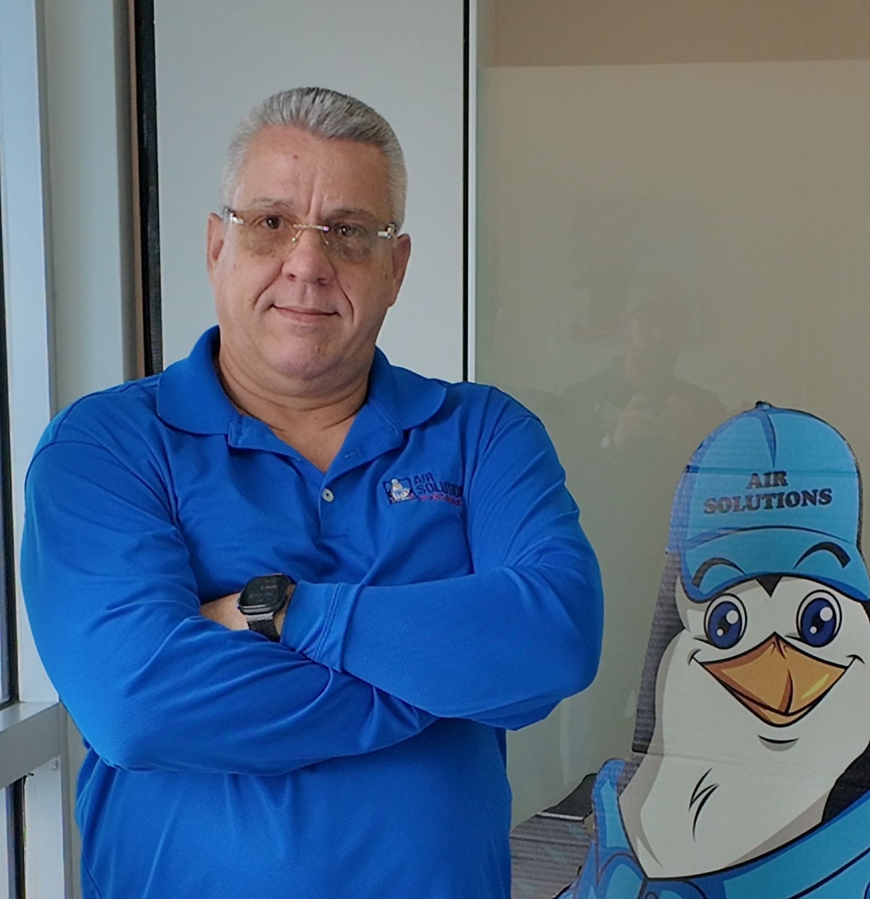 Man in a blue shirt with arms crossed stands beside a cartoon seagull sign indoors.