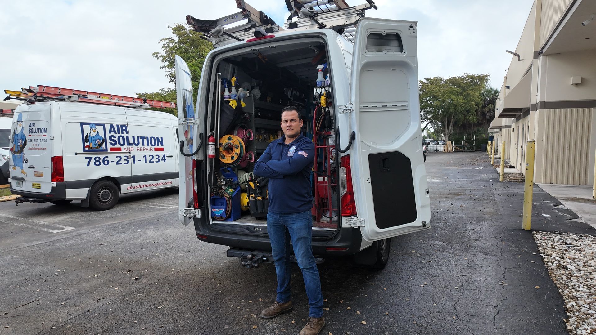 Worker standing beside an open white service van with tools in the back, in an industrial parking lot.