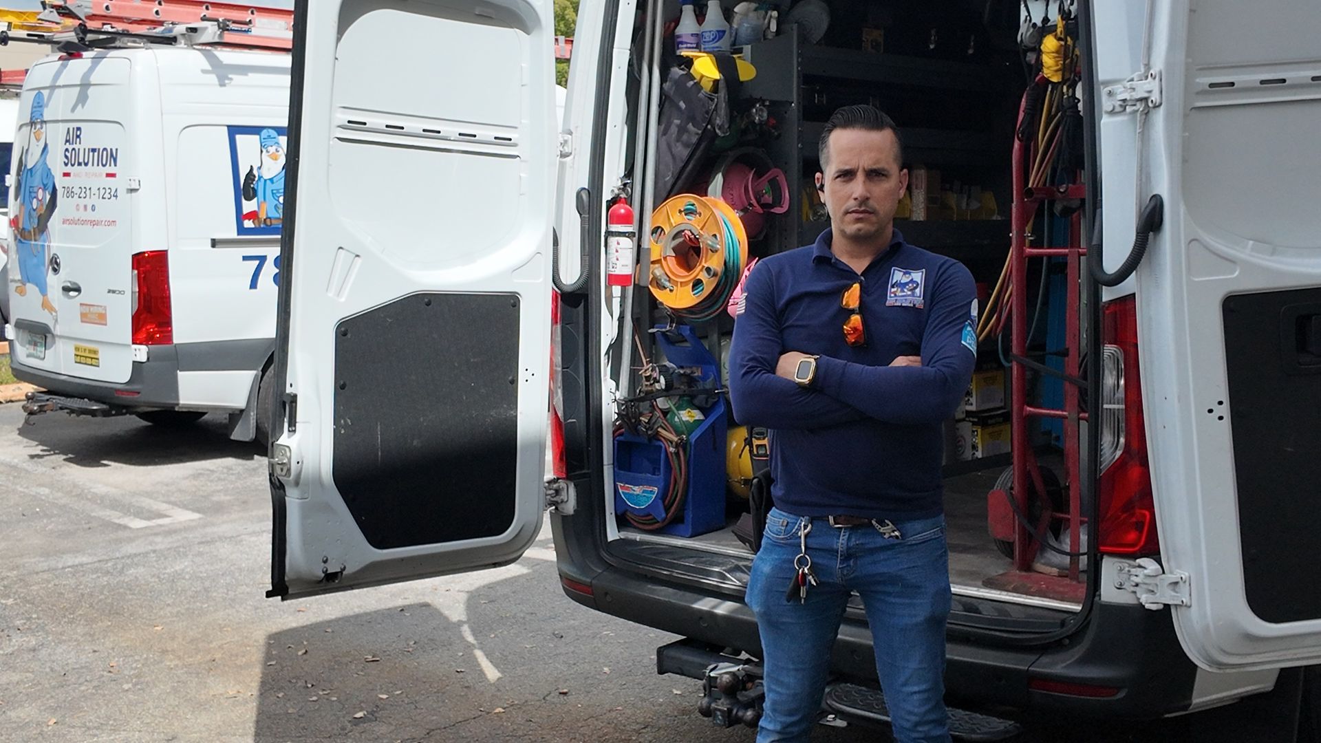 Man standing with arms crossed beside an open white utility van filled with equipment and hoses