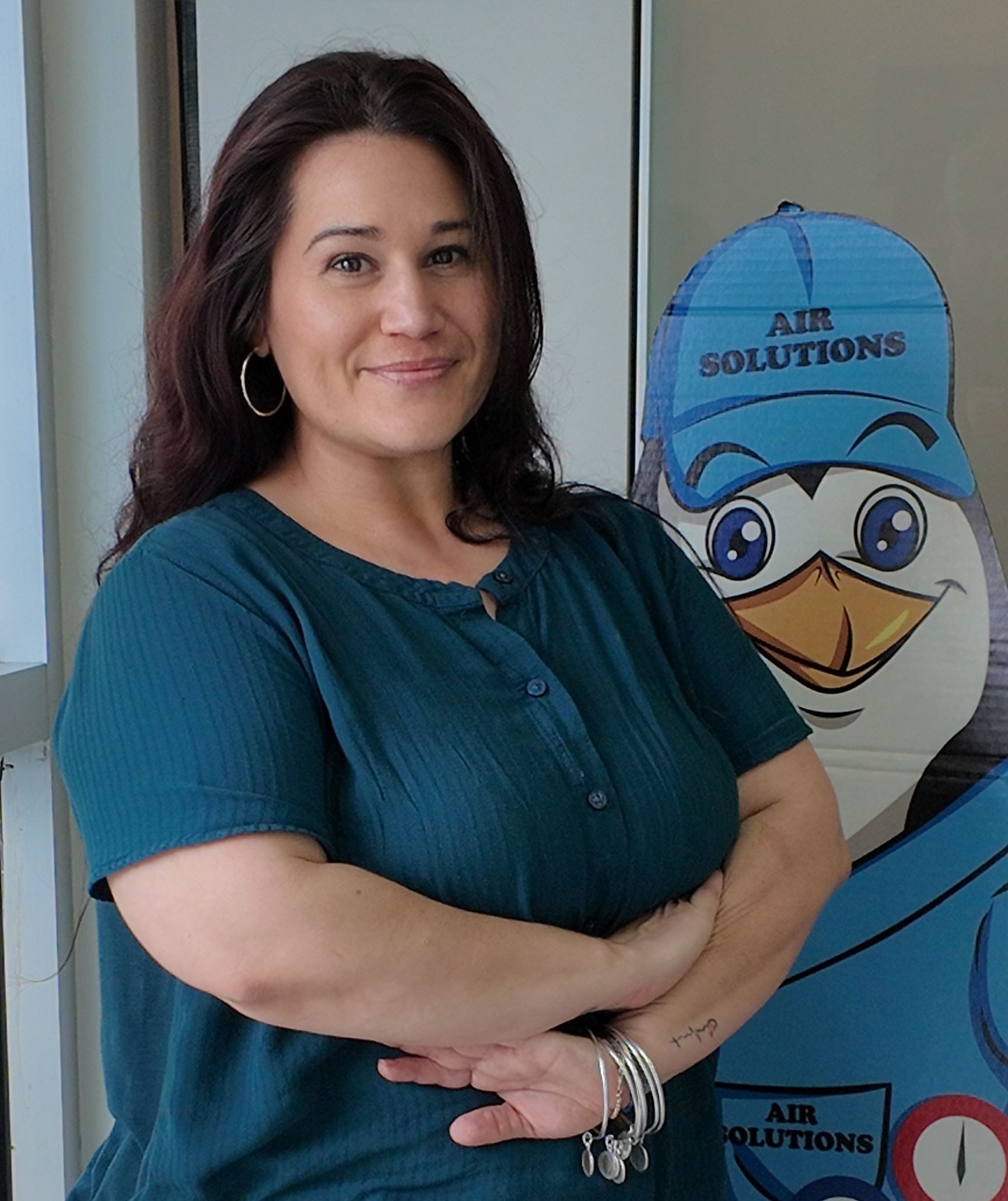Woman in teal shirt standing by a blue cartoon bird sign, arms crossed and smiling.