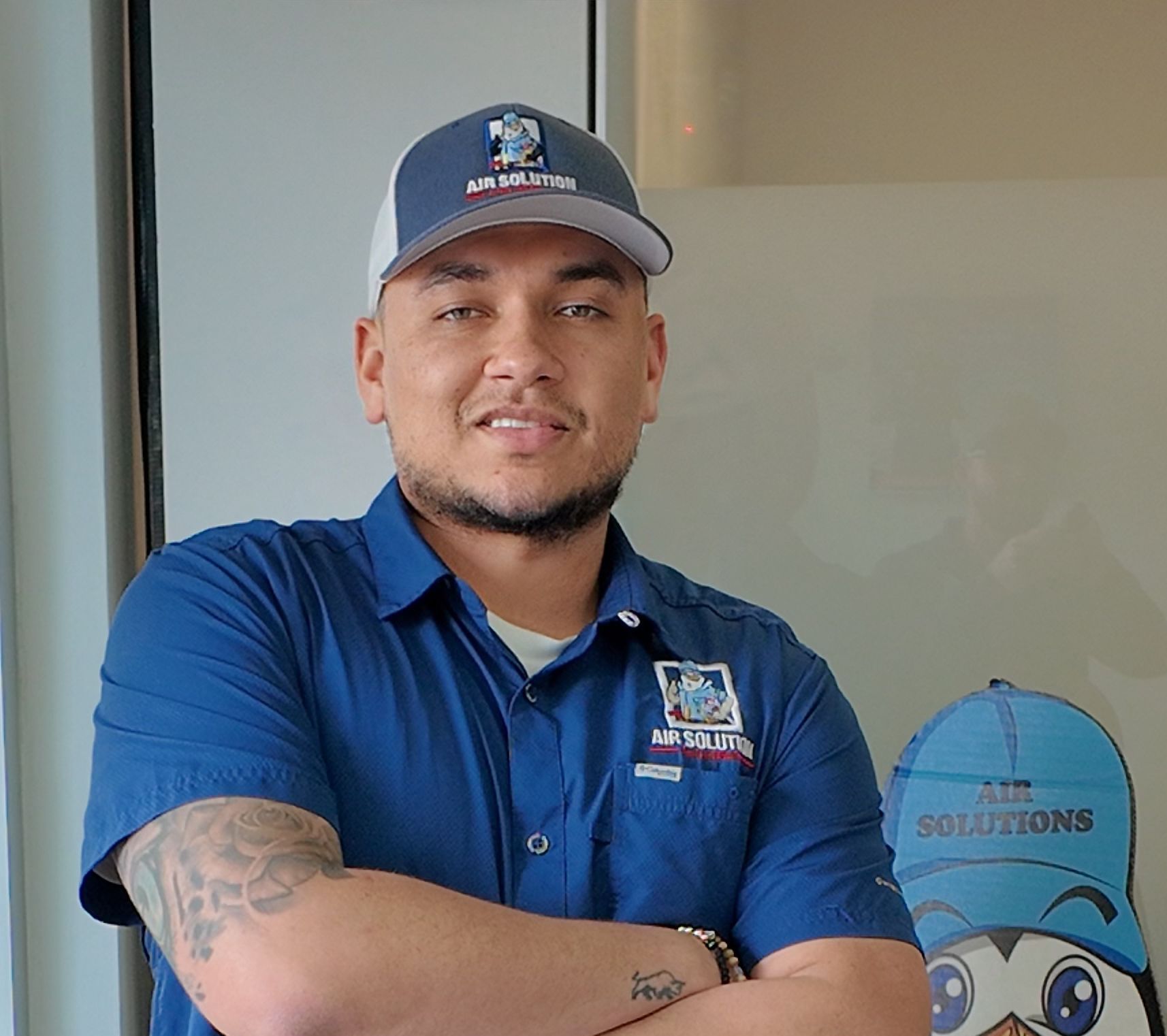 Man in blue shirt and cap standing with arms crossed beside a glass wall with a solar-themed sign