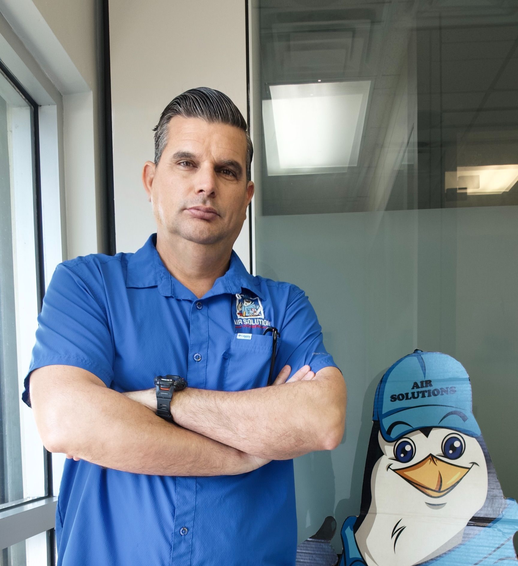 Man in blue polo stands with arms crossed beside a cartoon penguin in an office hallway.