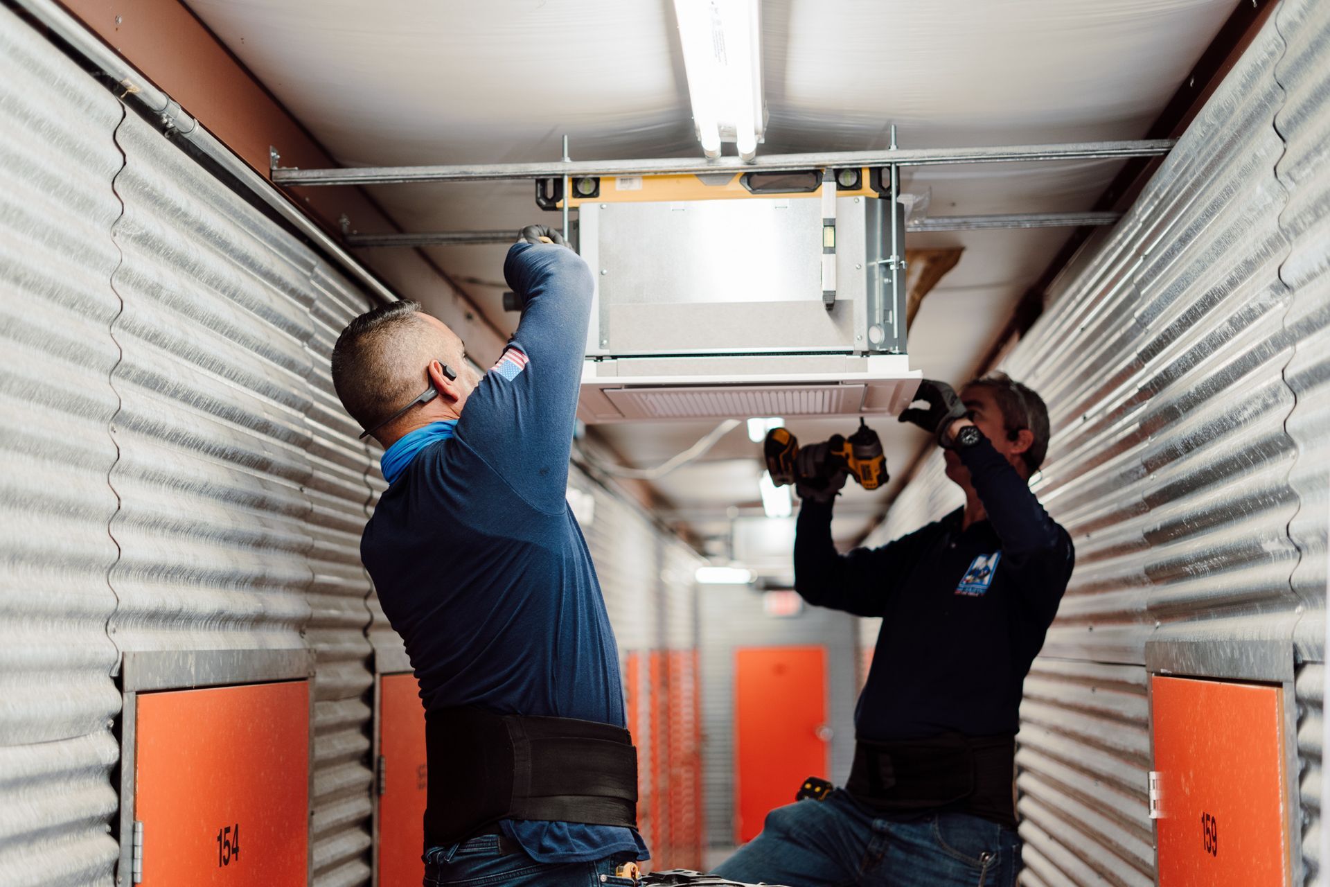 Two technicians installing a ceiling unit inside a narrow industrial corridor.