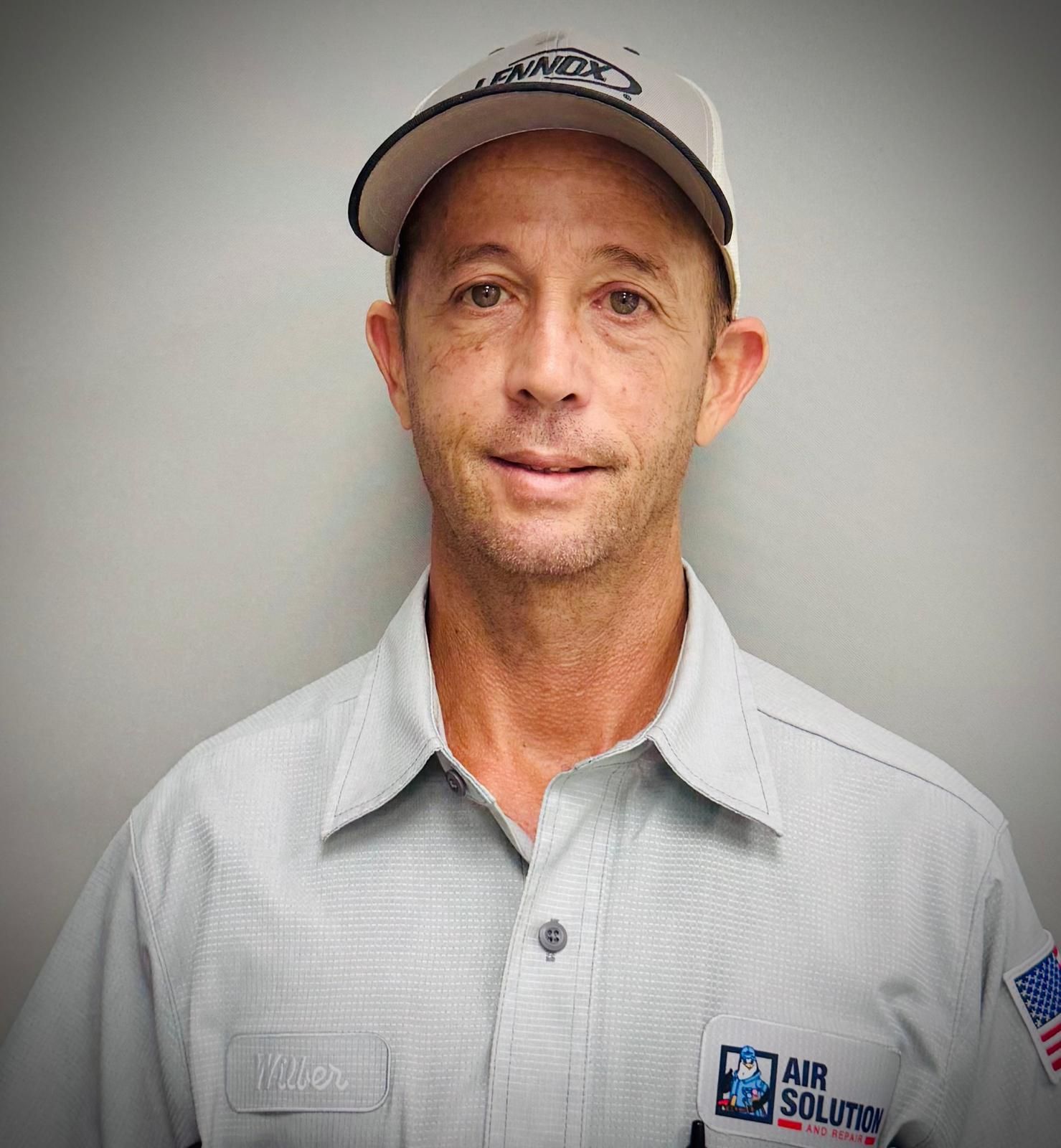 Man in a gray uniform and cap standing against a plain wall, facing the camera.