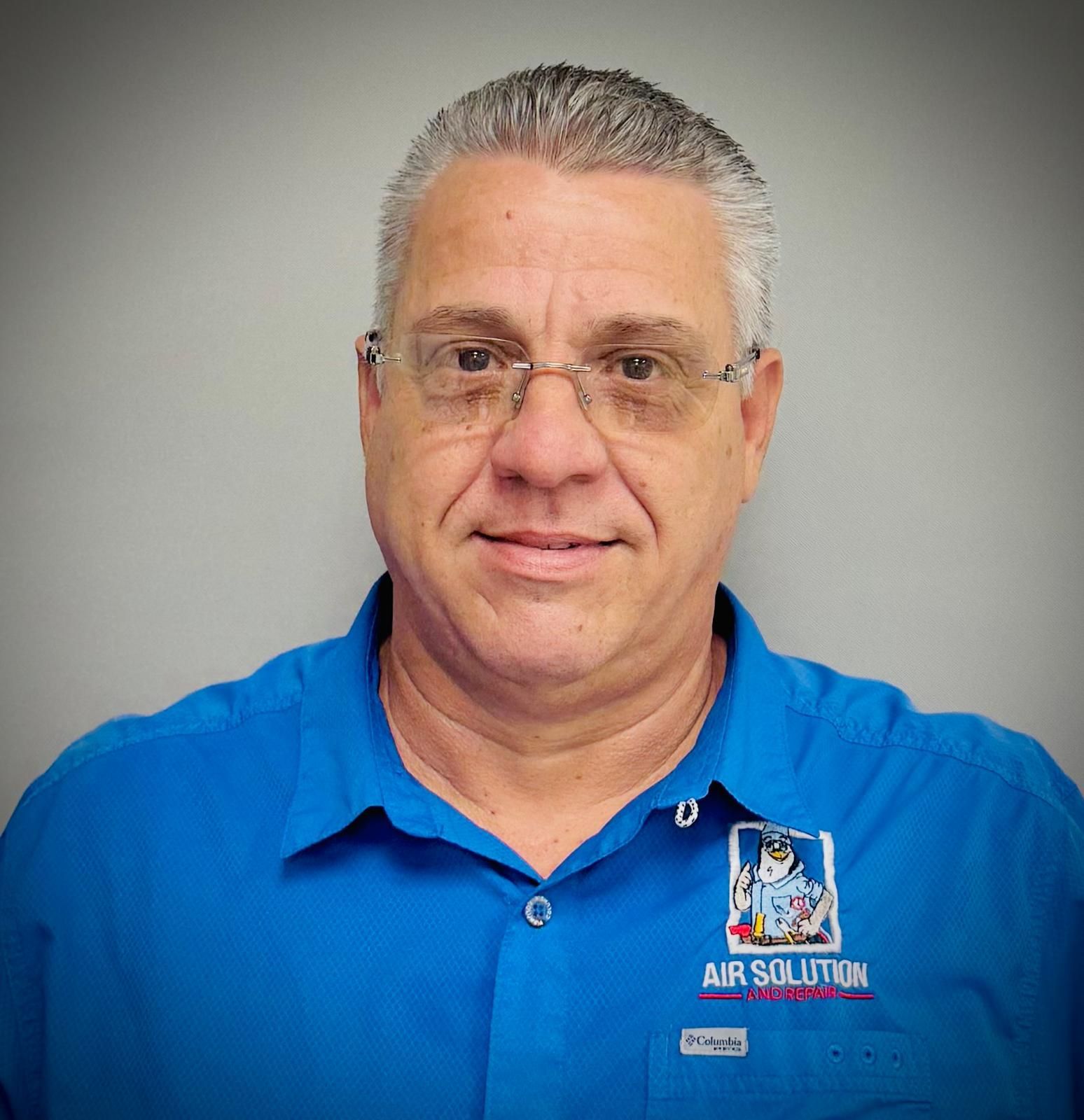 Portrait of a man in glasses wearing a bright blue polo shirt with a logo on the chest, against a gray background