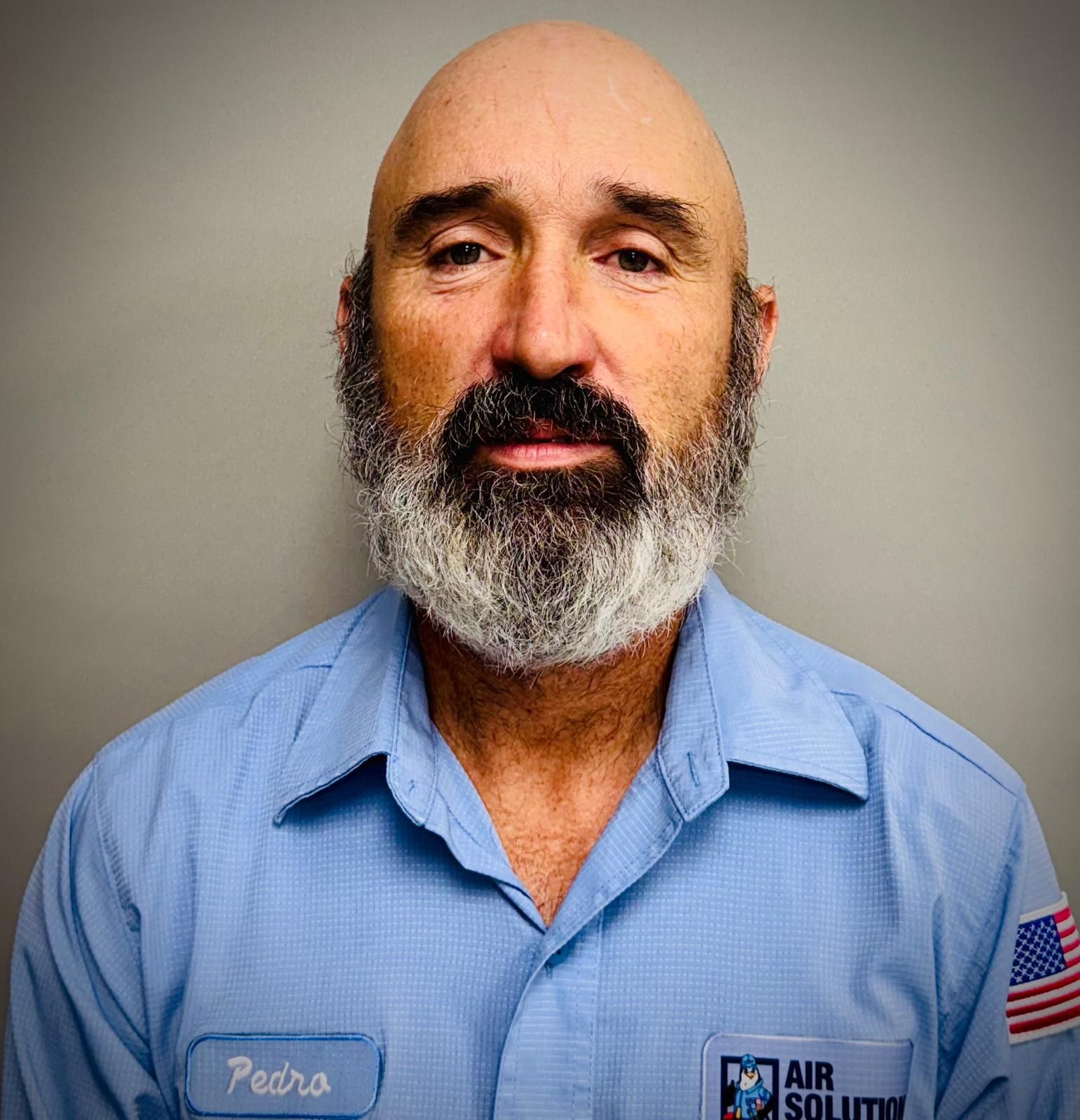 Portrait of a bearded man in a blue work shirt with an Air Force patch against a plain background