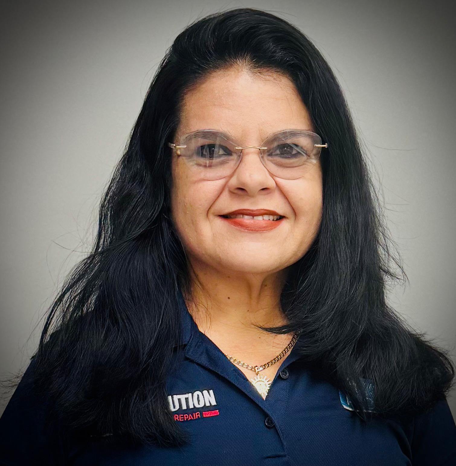 Portrait of a smiling person with long dark hair, glasses, and a navy shirt with a company logo on a gray background