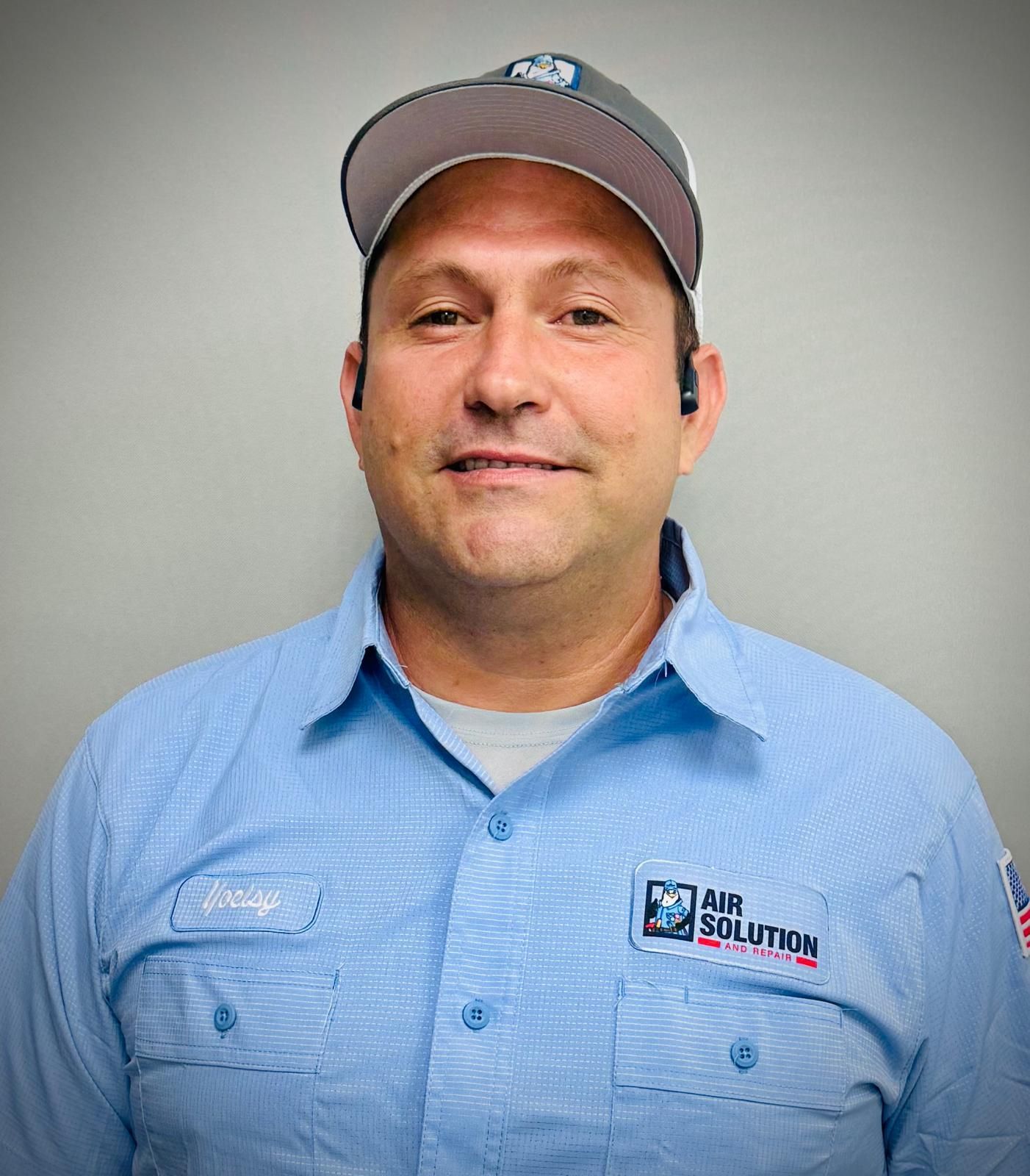 Man in a gray cap and blue work shirt smiling against a plain gray background