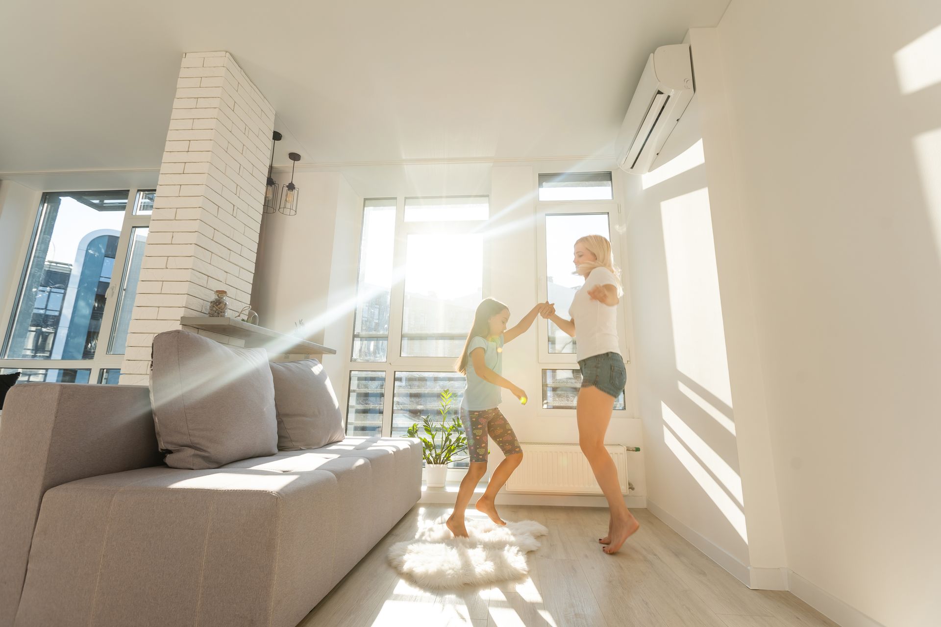 Two people dancing in a bright sunlit living room with a couch, windows, and wall-mounted air conditioner