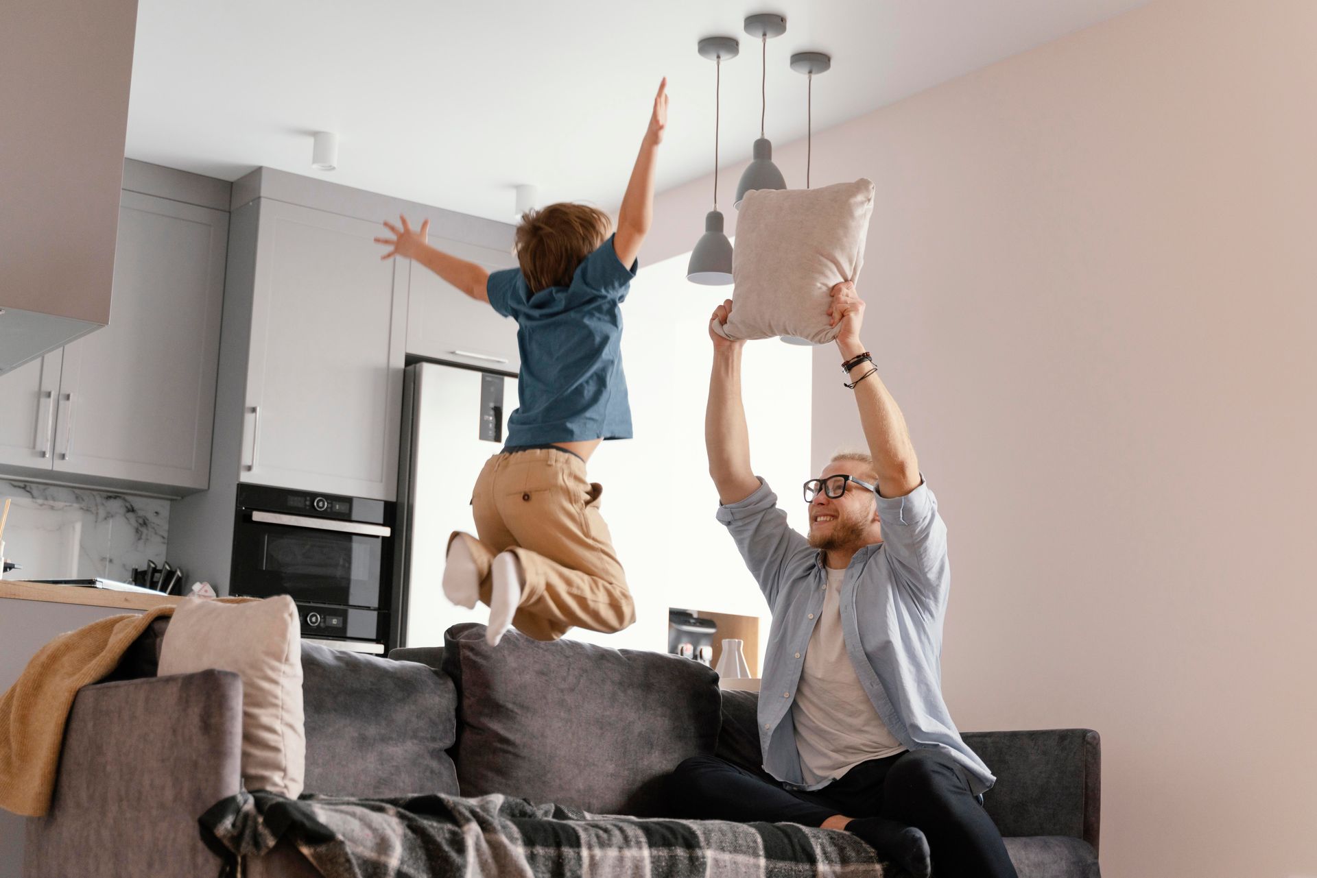 Child jumping on a couch as an adult lifts a pillow in a bright living room