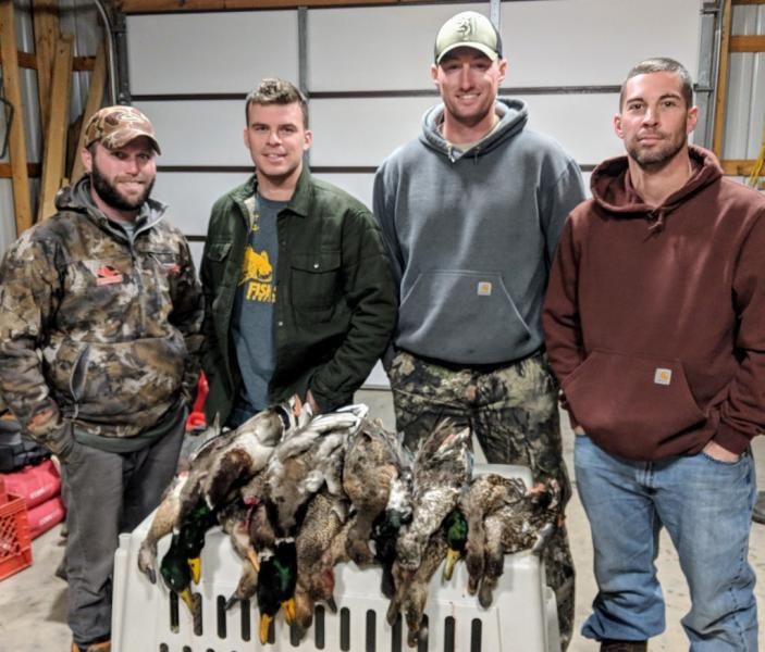Four men pose indoors behind a crate of hunted ducks and geese, wearing camo and casual clothes.