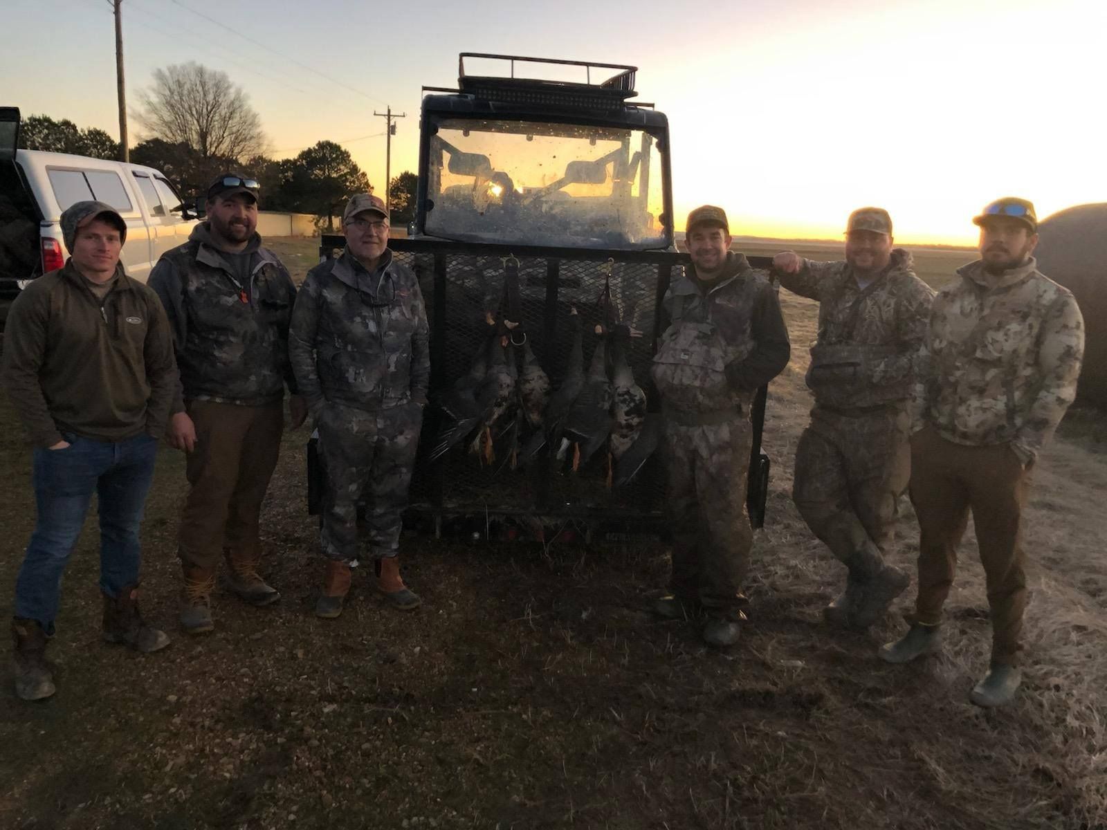 Six people stand beside a muddy utility vehicle at sunset on a dirt road.