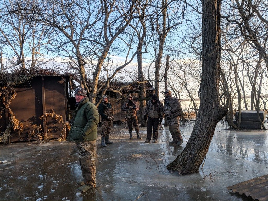 People in winter clothing stand on a frosty path near a small wooden shelter among bare trees.