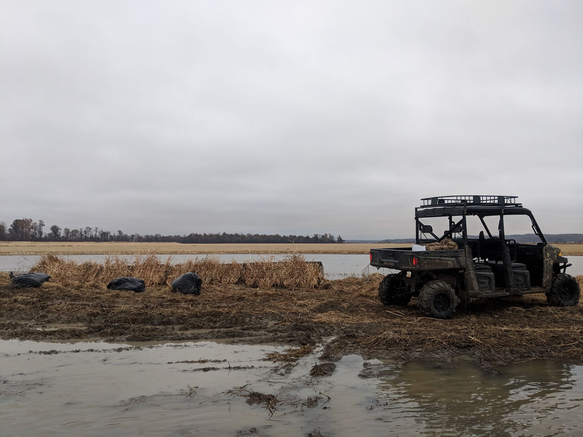 Muddy farm field with a black utility vehicle near a flooded ditch under a gray sky
