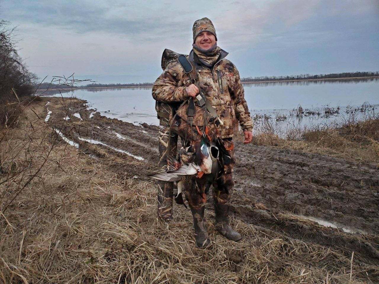 Camouflaged person standing outdoors by a snowy wetland, holding several hunted ducks