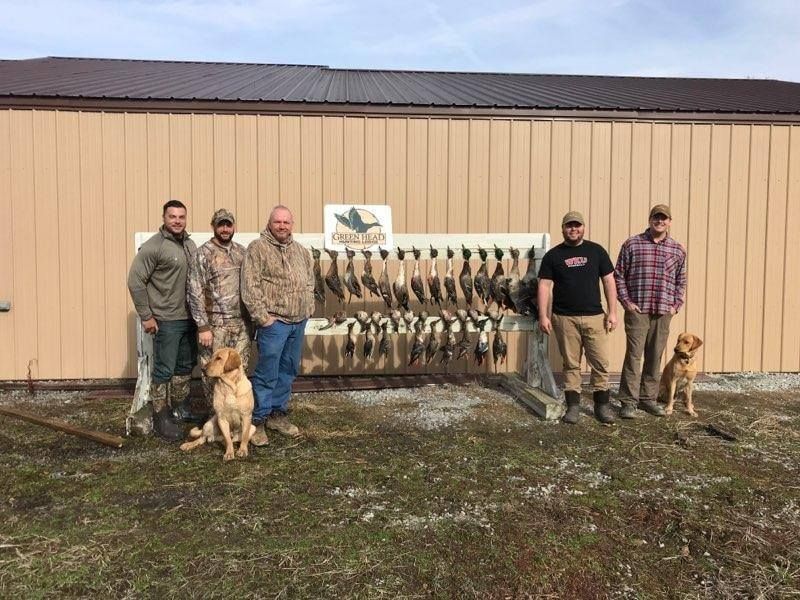 Five people with a dog stand before a wall of hanging game birds outside a building.