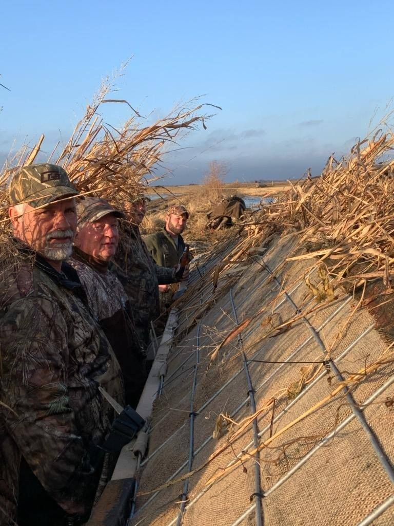 Hunters in camouflage stand beside a reed blind in a marsh at dawn