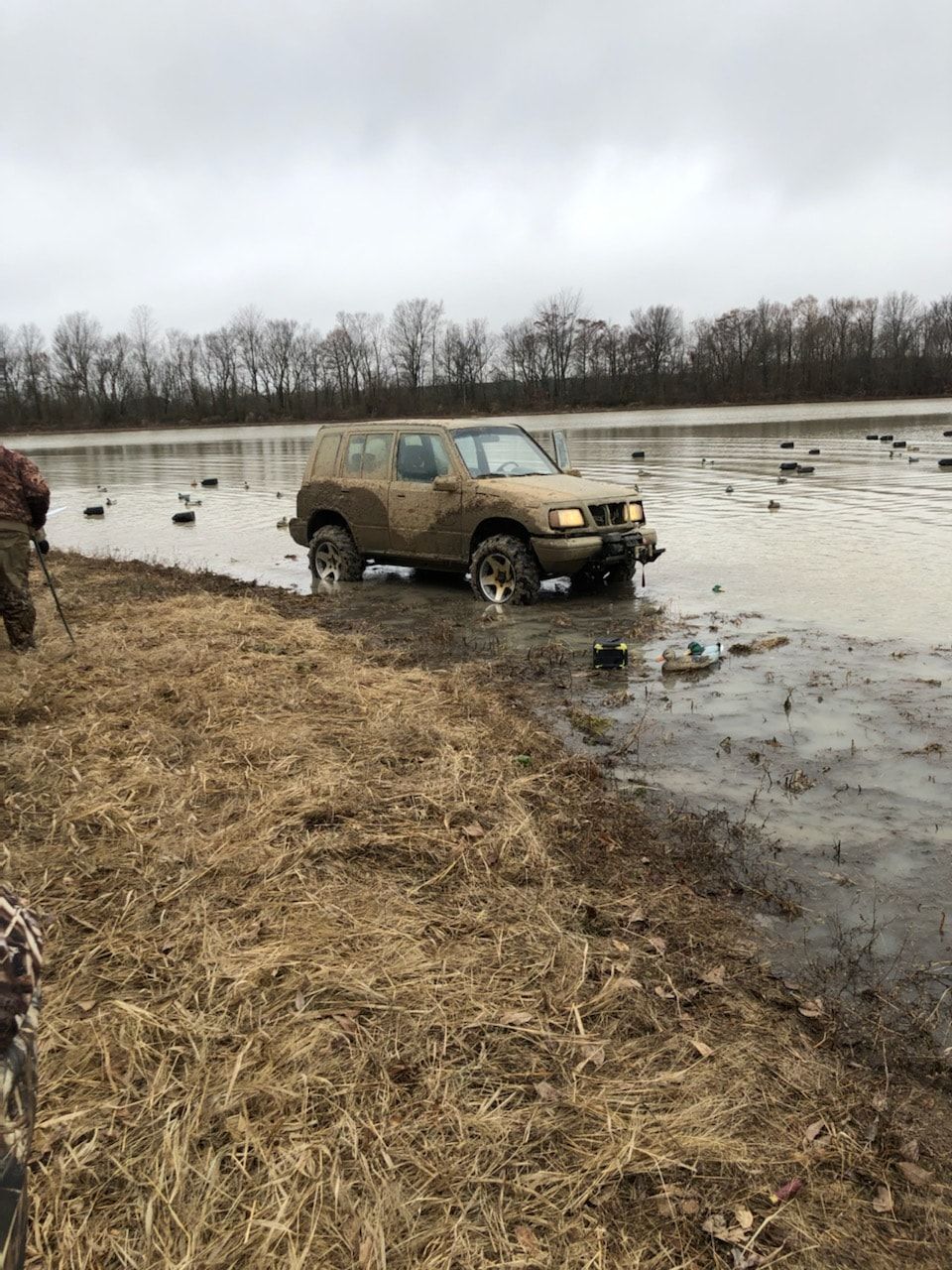 Mud-covered SUV driving through a flooded field near a grassy bank under a gray sky