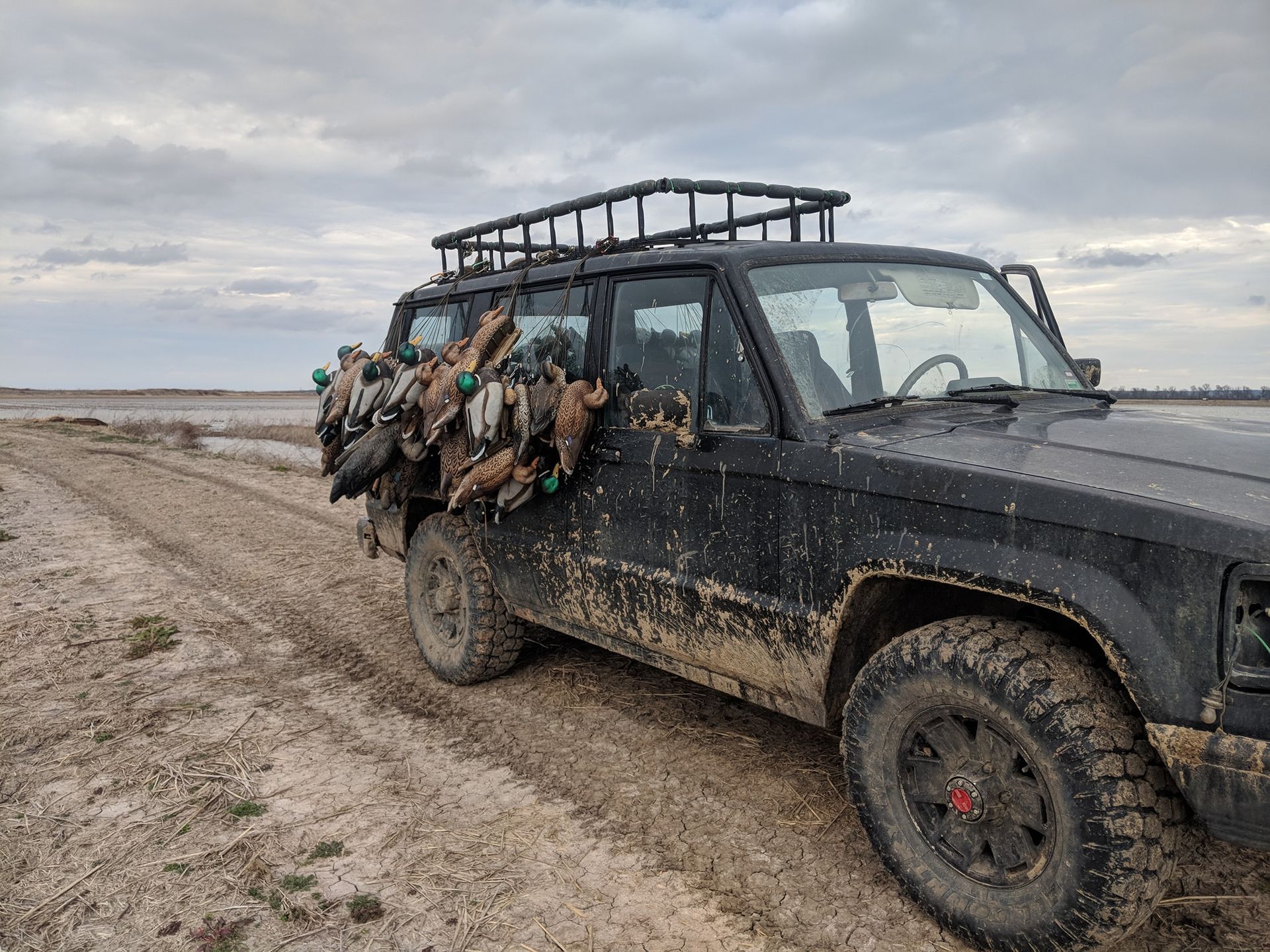 Mud-covered black off-road truck on a dirt track, loaded with gear under a cloudy sky.