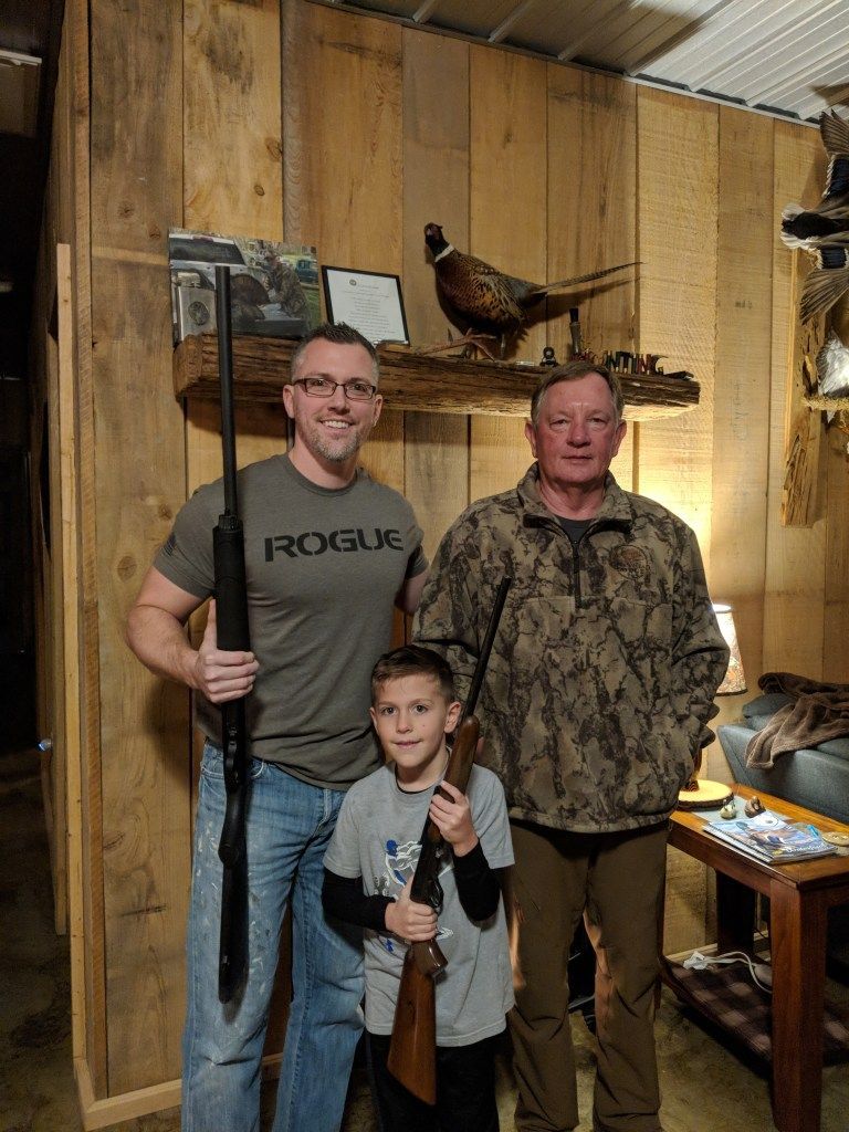 Three people standing indoors with a mounted turkey and a hunting rifle against wood-paneled walls.