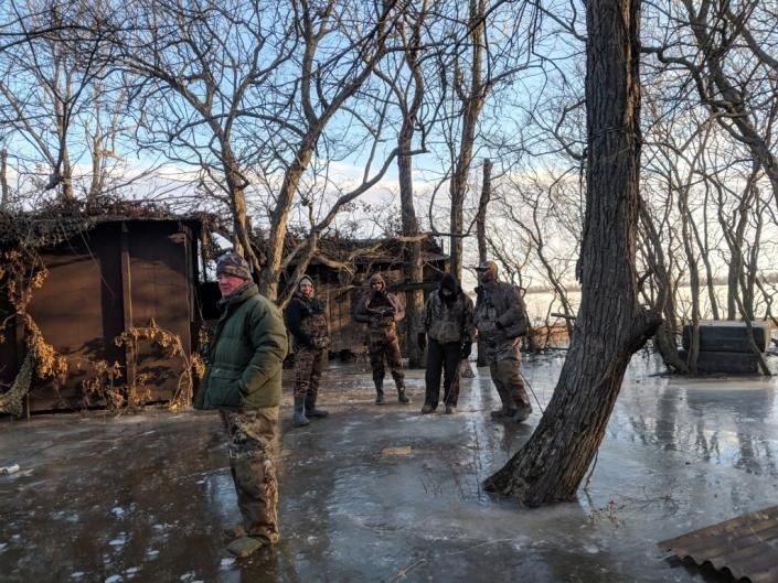 People standing on icy ground near a woodpile and shed in a winter wooded area.
