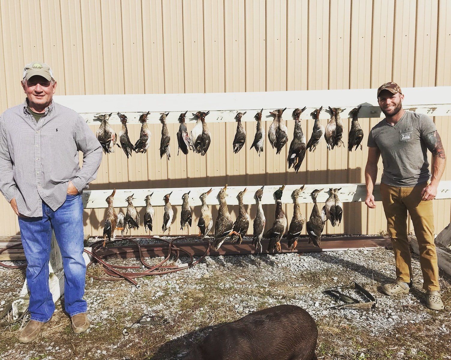 Two men standing beside rows of hanging game on racks outdoors by a wooden fence.