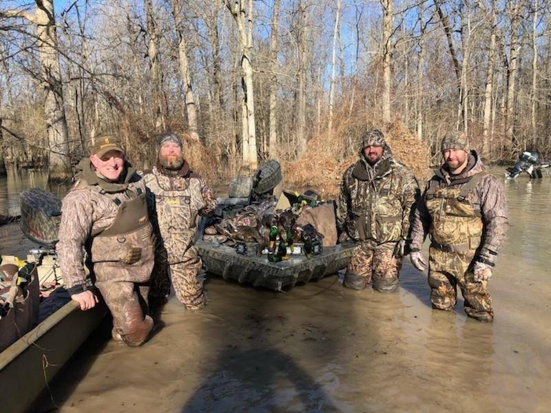 Five people in camouflage stand in shallow muddy water beside an all-terrain vehicle in a bare woodland.