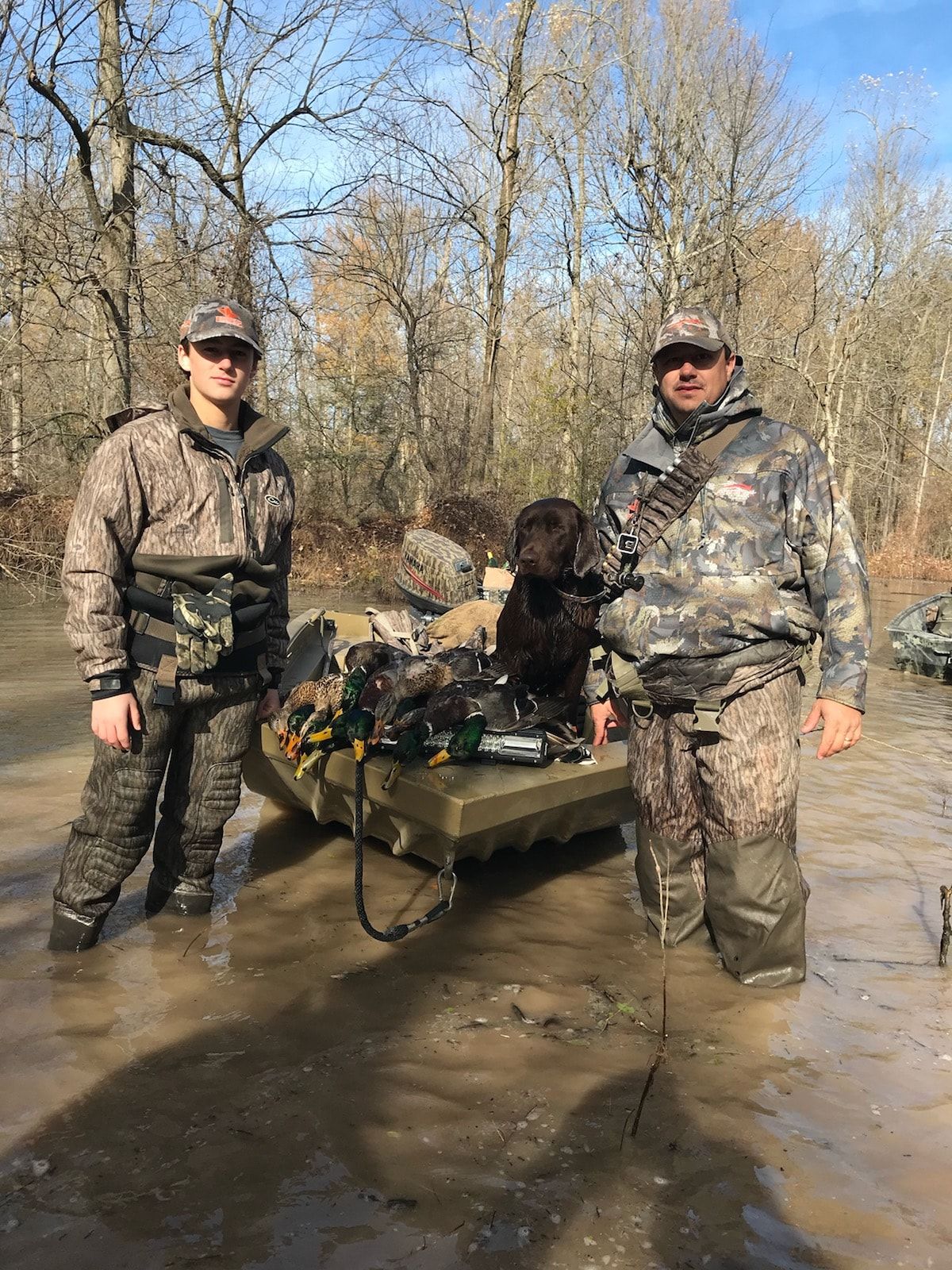Two hunters stand by a camouflaged cart with a black dog in a muddy wooded area.